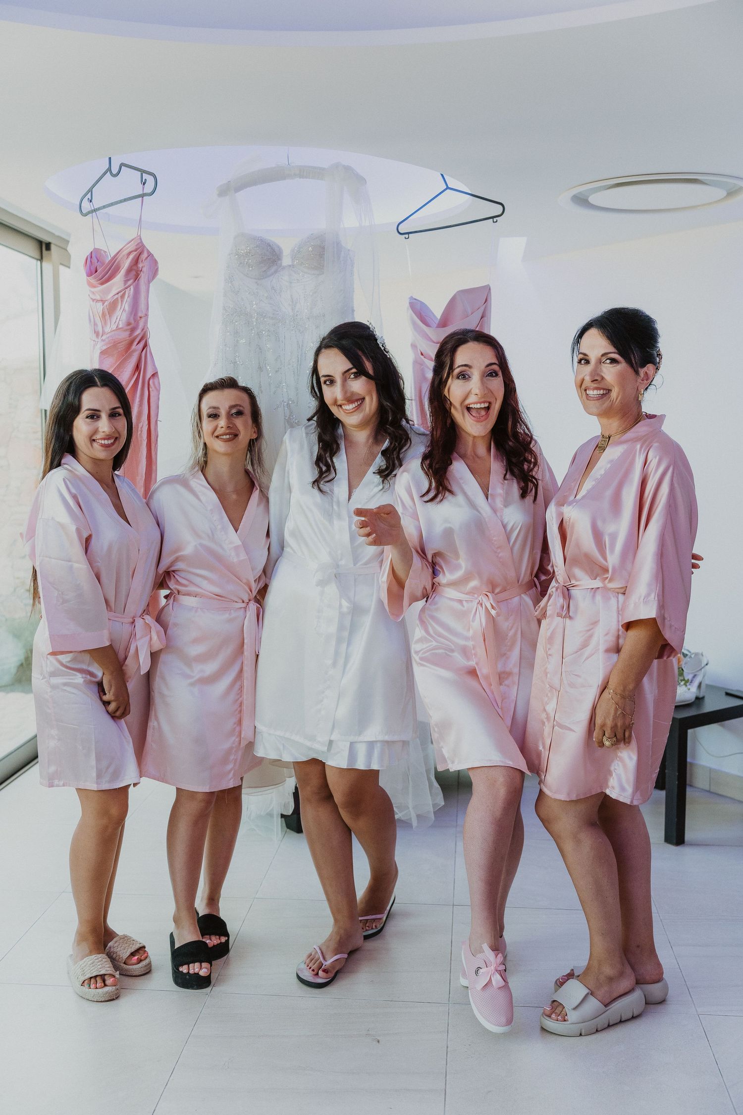 Bridal party members wearing pink robes stand together in a dressing room with a wedding gown hanging behind them.