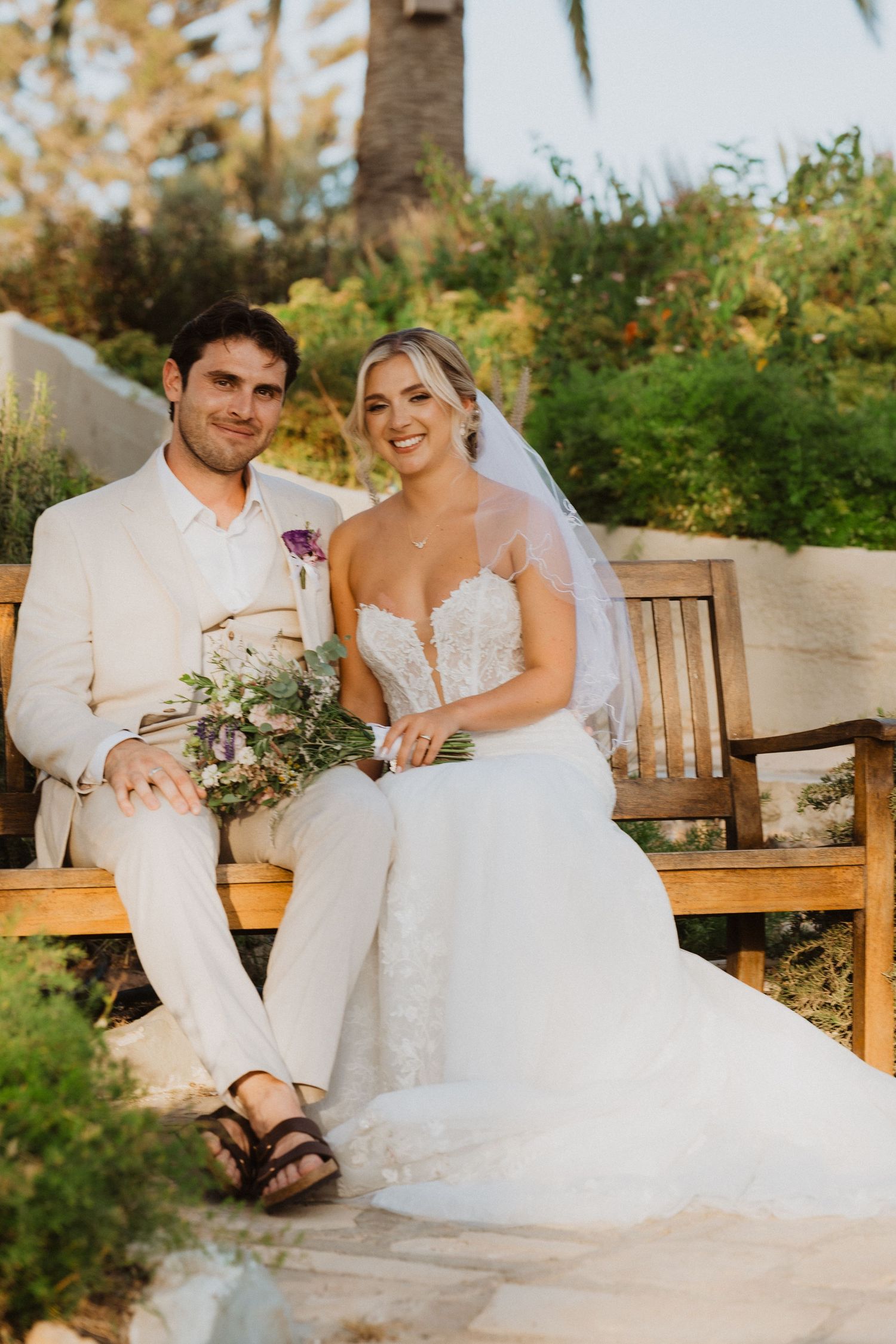 Couple in wedding attire shares candid laughter while relaxing on a garden bench.