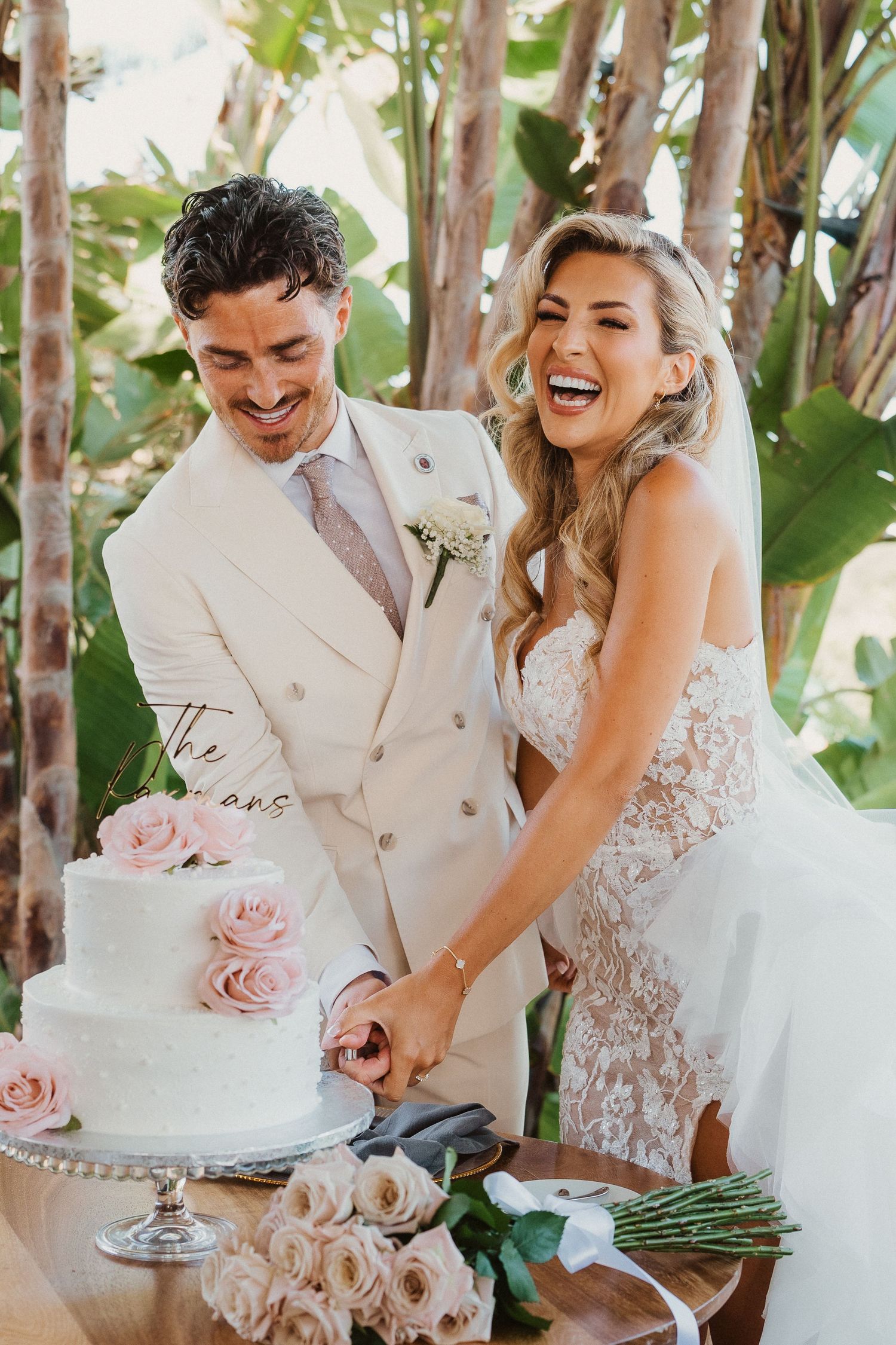 Bride and groom share joyful moments cutting their elegant white wedding cake decorated with pink roses in outdoor setting.