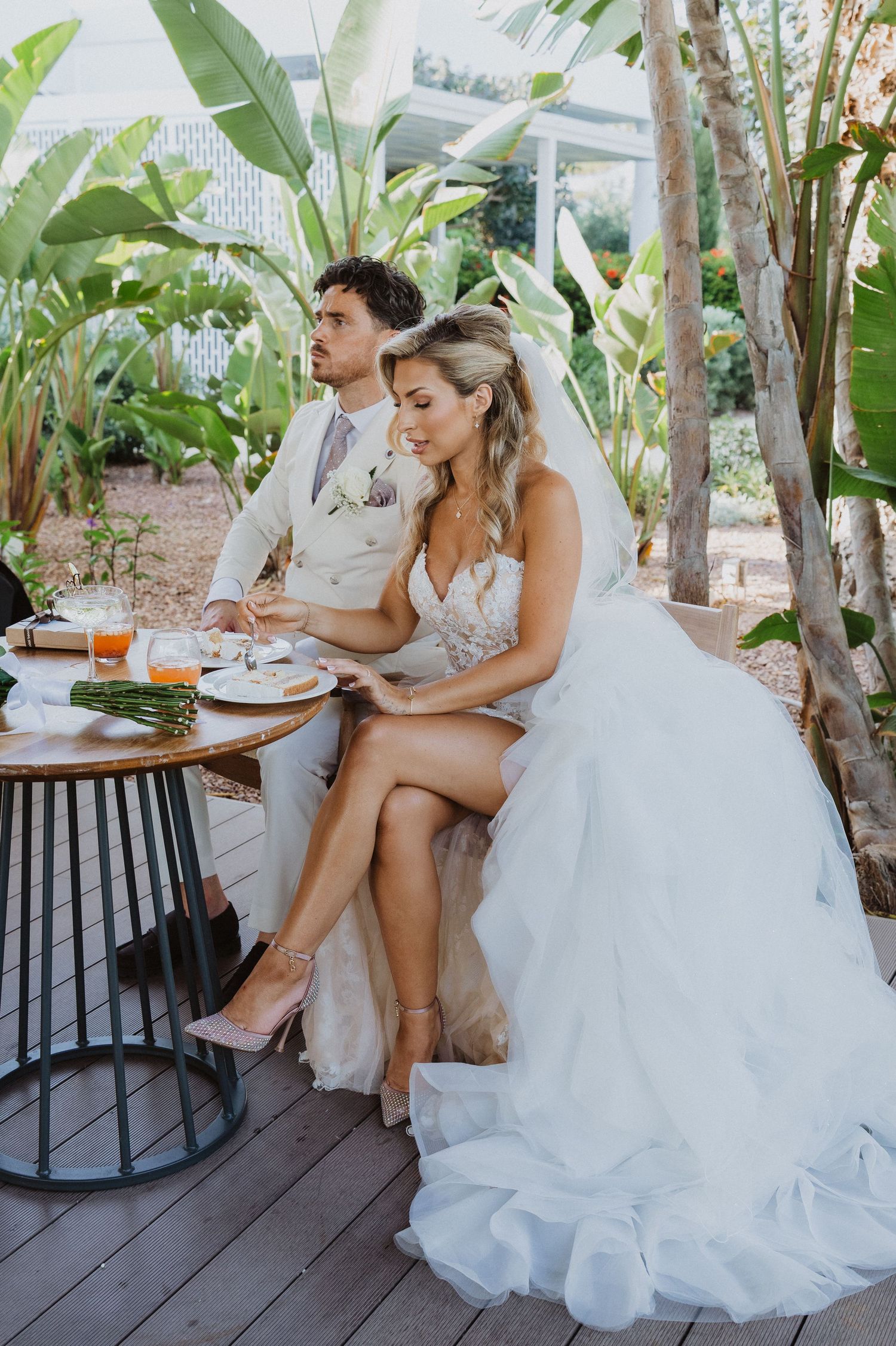 A couple sits together at a rustic outdoor table surrounded by tropical plants on a wooden deck.