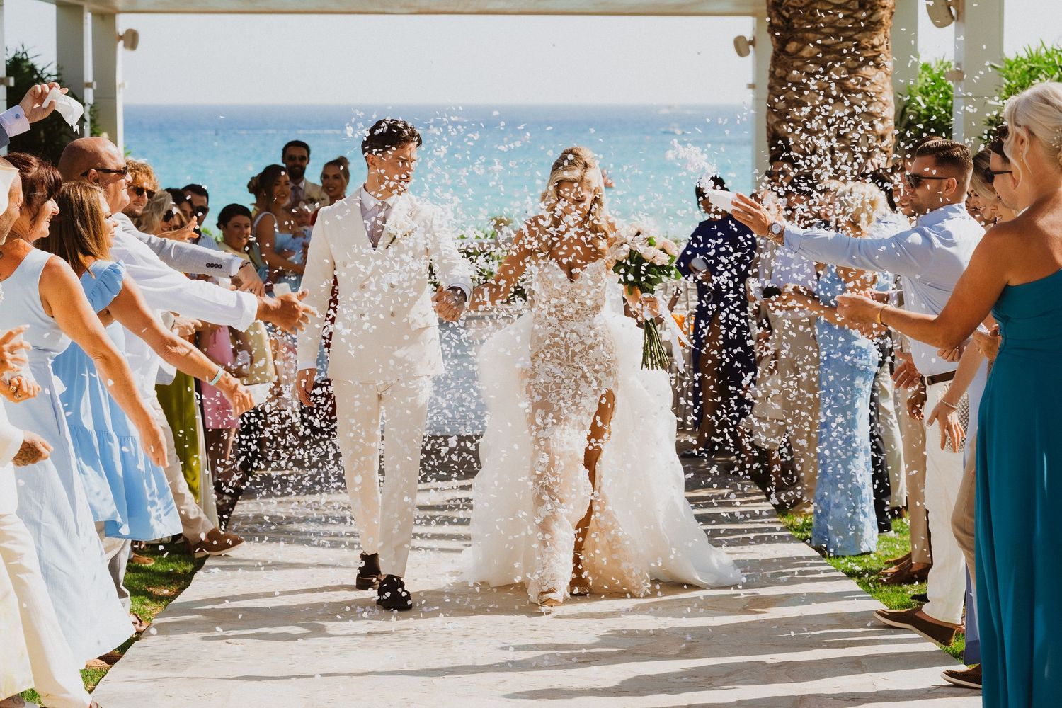 Wedding guests throw flower petals as newlyweds walk down an outdoor aisle by the ocean.