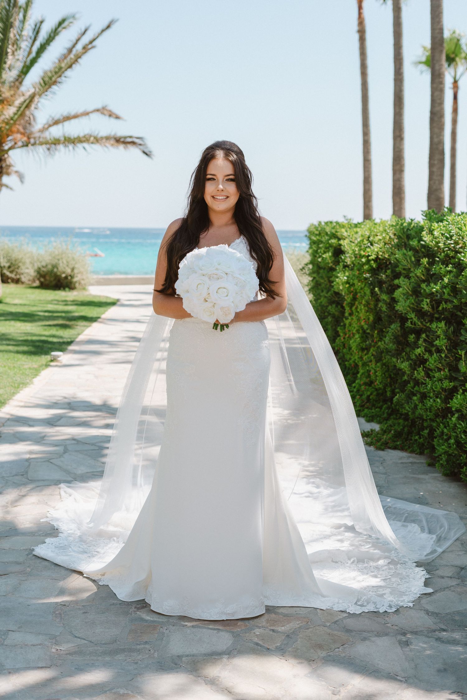 Bride in flowing white wedding gown poses along palm-lined coastal pathway.