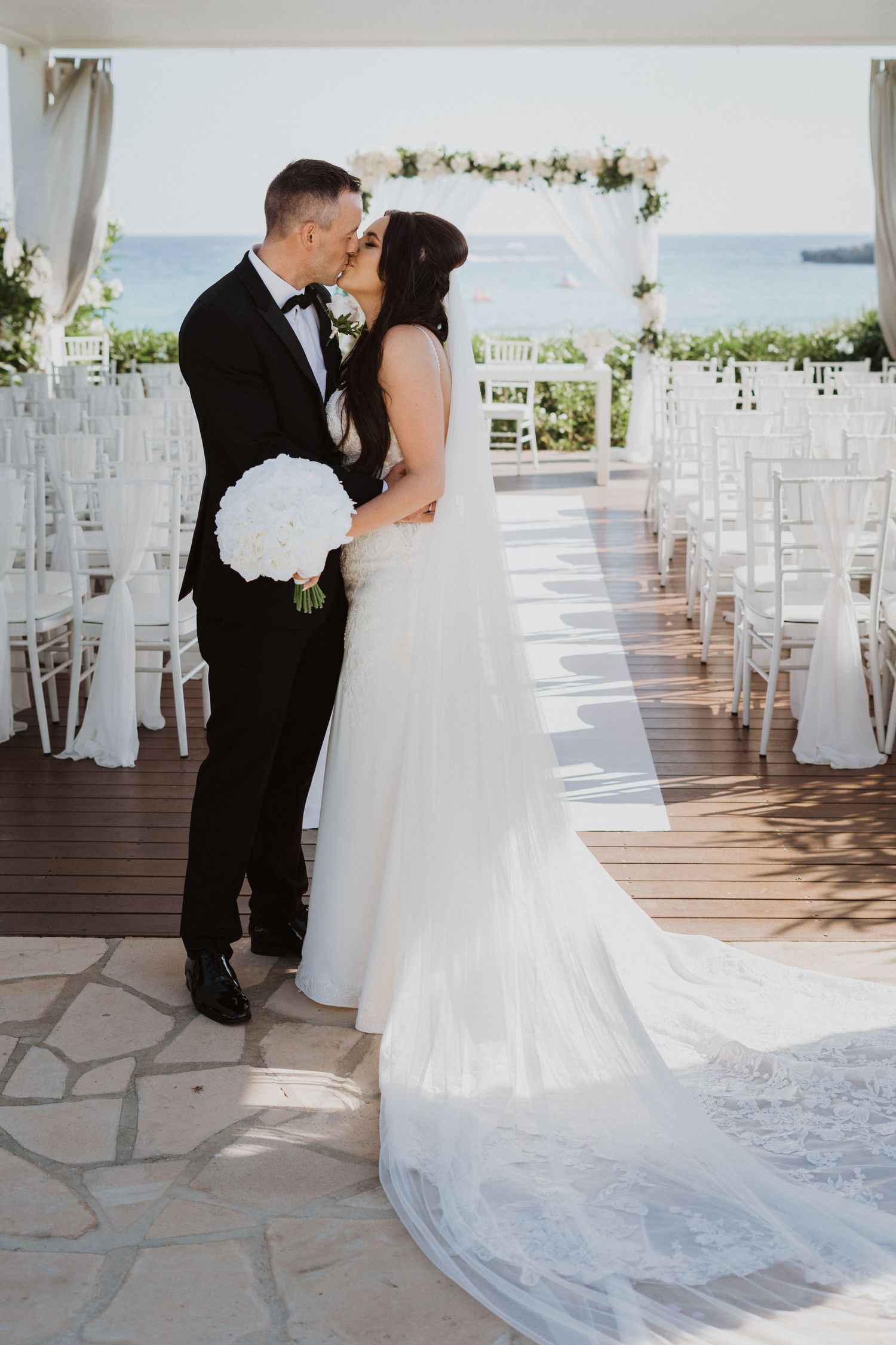 Wedding couple poses on wooden deck with white ceremony chairs in background.