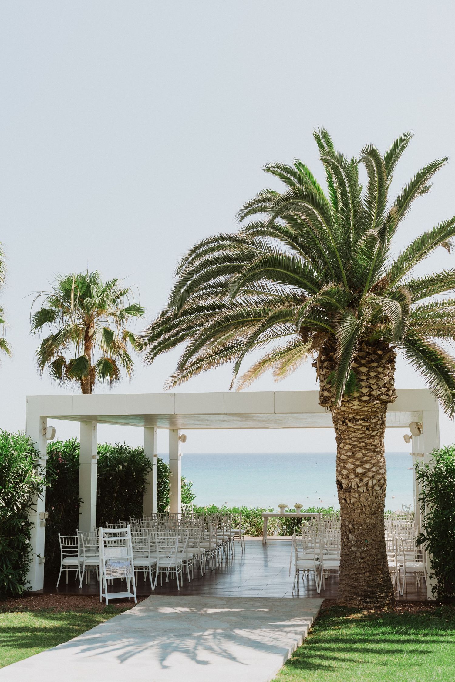 Palm trees line a white-columned beachfront wedding venue with chairs set up for a ceremony overlooking the ocean.