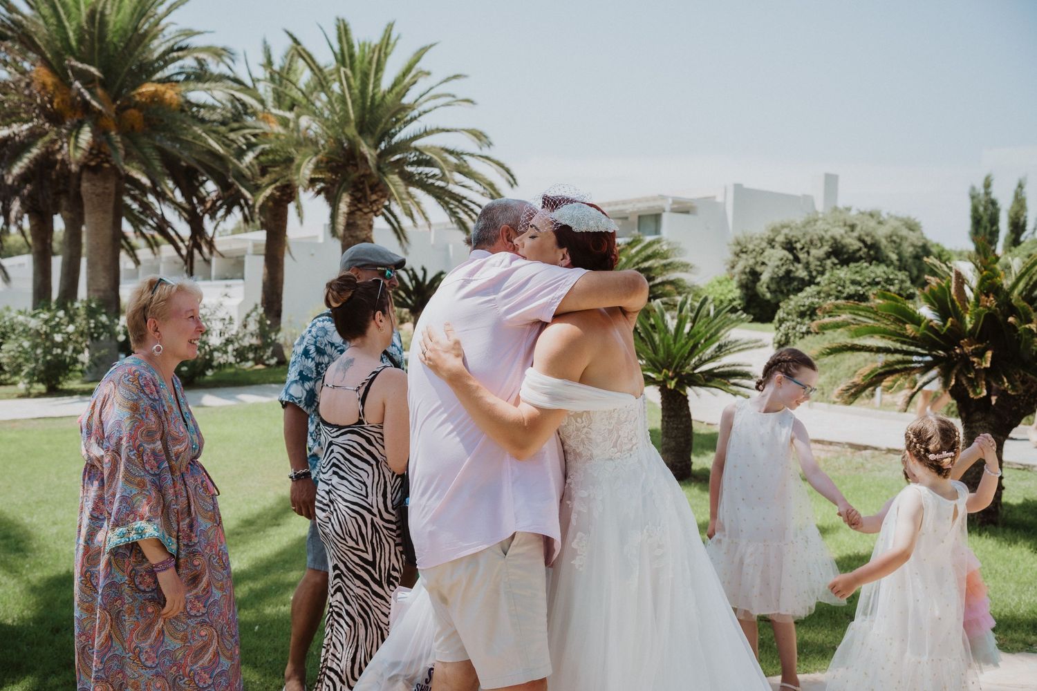 Wedding celebration with guests gathering for hugs on a sunny palm-lined pathway.
