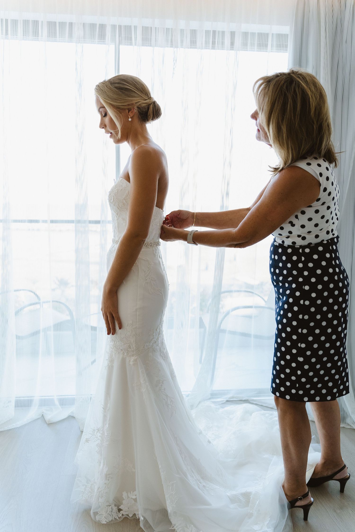 A wedding dress fitting session with final adjustments being made near a bright window.