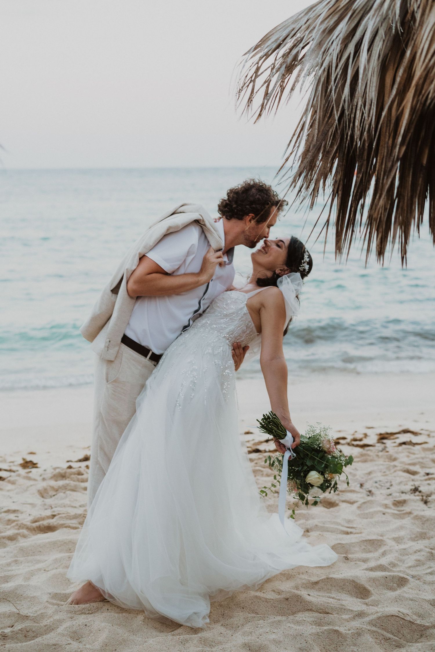 Romantic dip pose wedding photo on sandy beach with palm fronds in frame.
