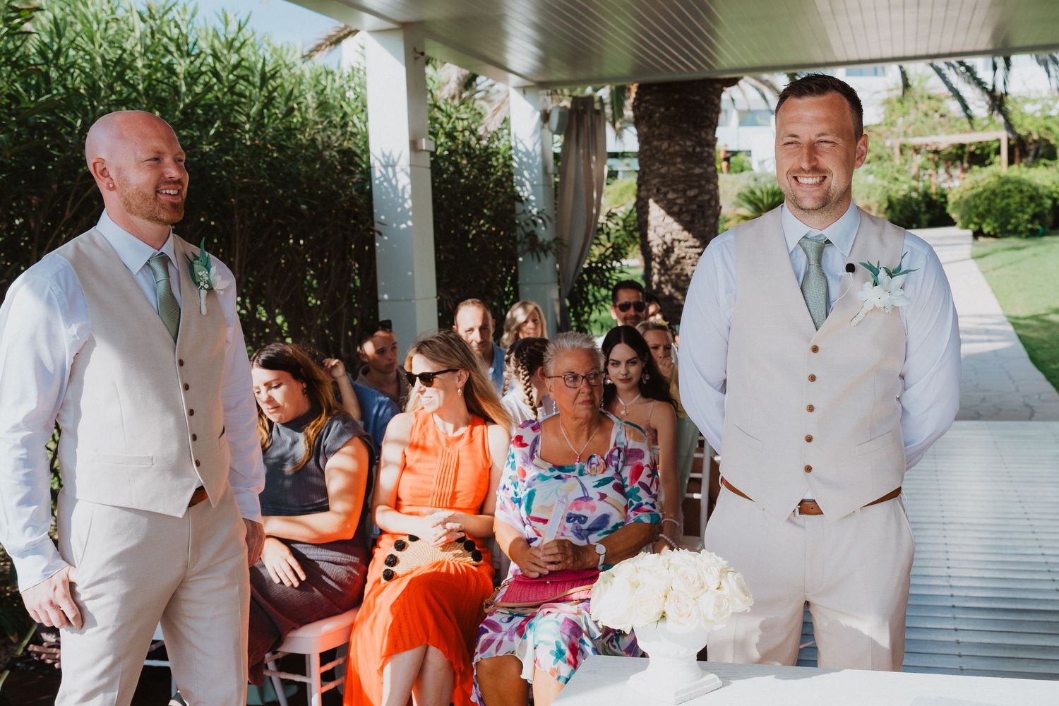 Wedding guests in orange and floral attire gather under a covered patio area during an outdoor ceremony.