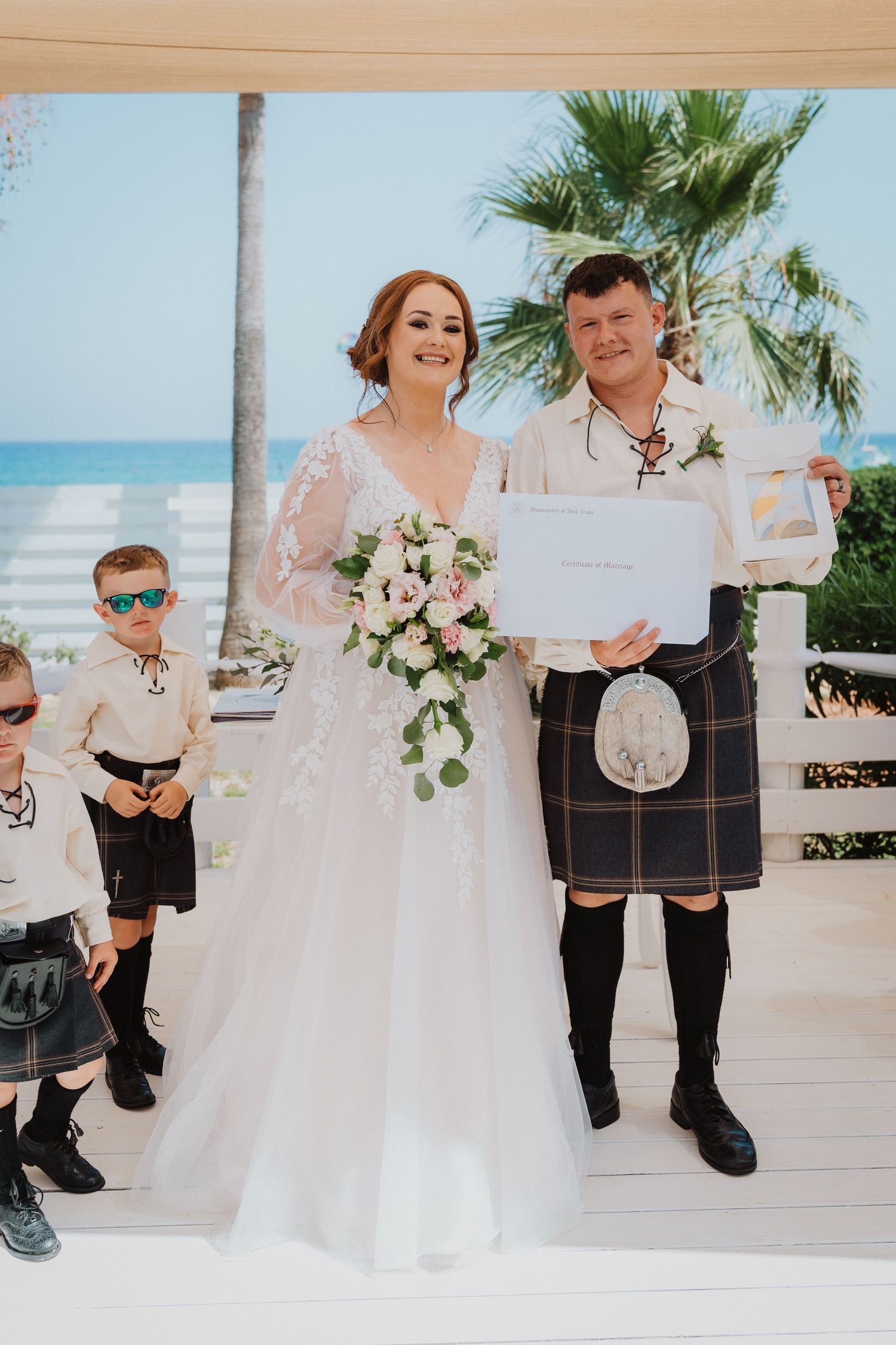 Wedding couple displays marriage certificate at beach ceremony with children in Scottish attire.
