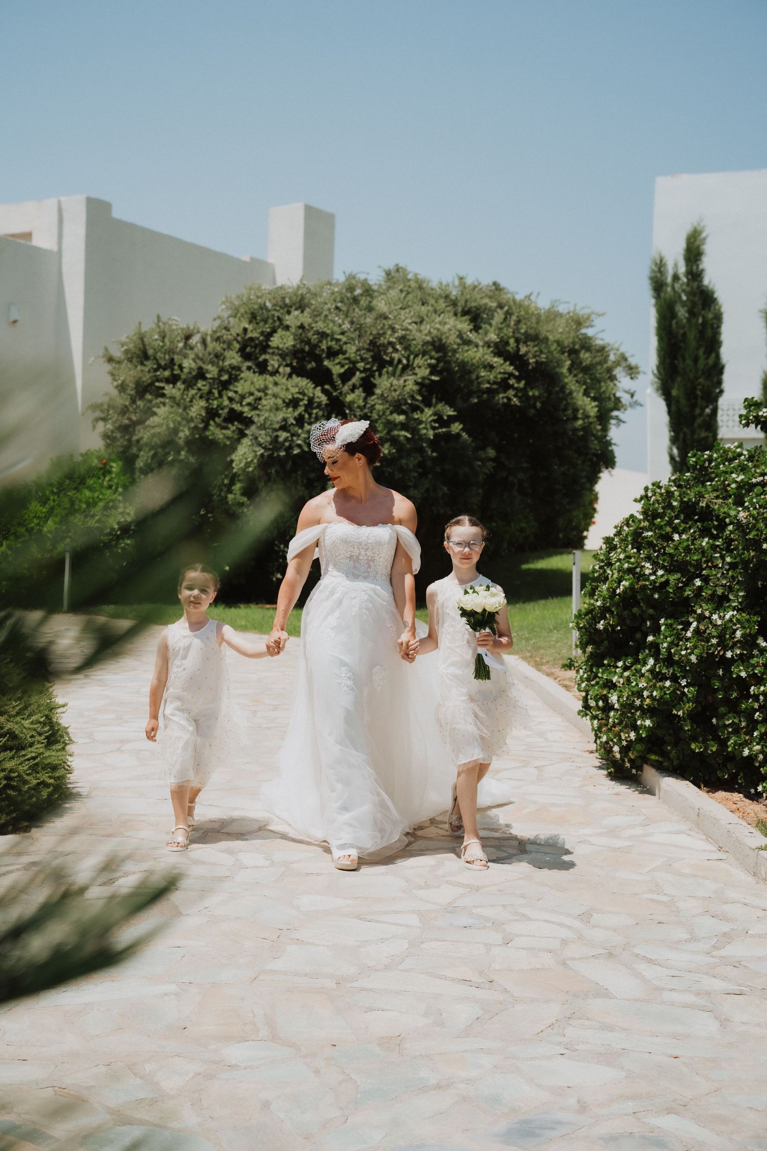 A bridal party walking along a stone path surrounded by lush greenery on a bright summer day.