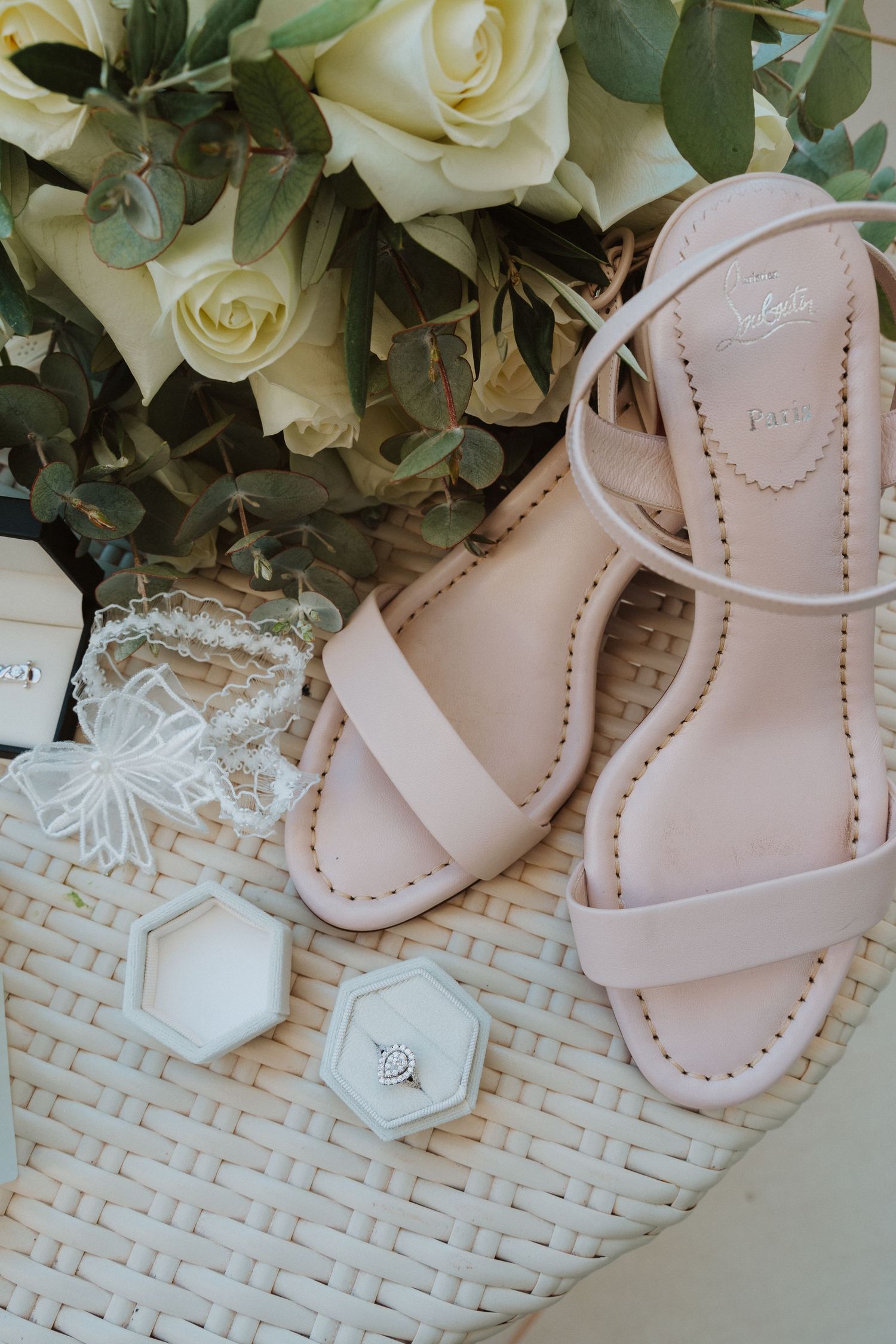 Elegant pale pink sandals and white roses arranged on a woven surface with hexagonal ring boxes.