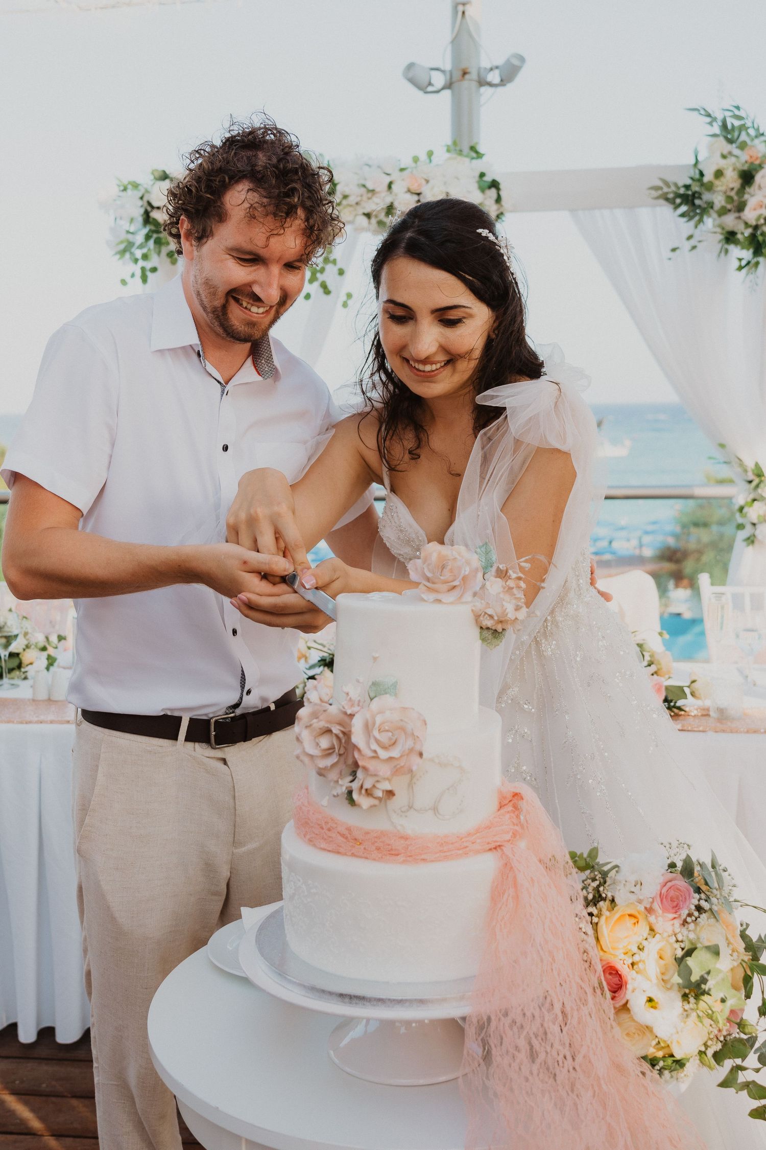 A couple enjoys cutting their elegant white wedding cake decorated with pink accents at an outdoor reception.