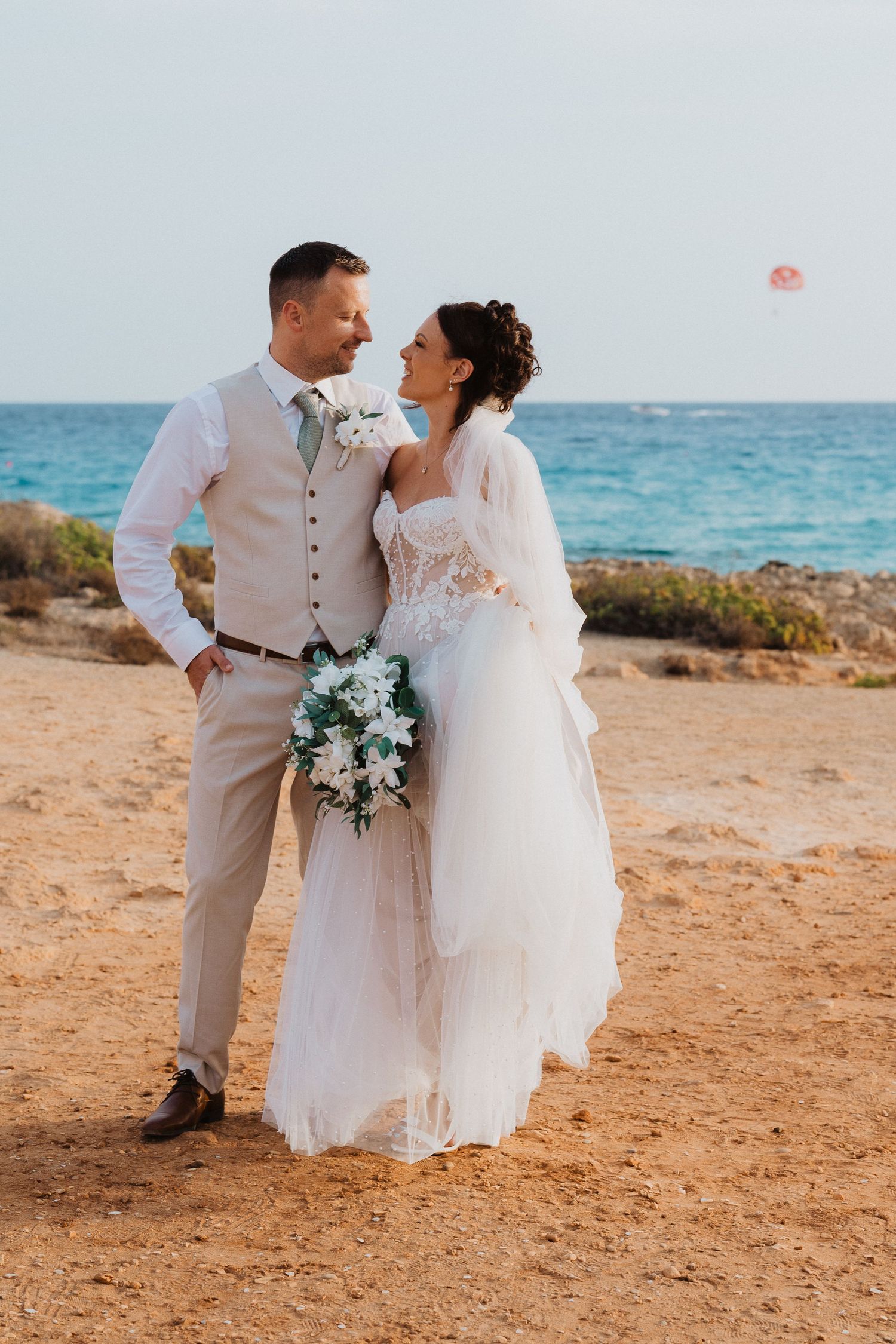 A romantic beach wedding scene with a couple in formal attire standing on sandy shores against blue ocean waters.