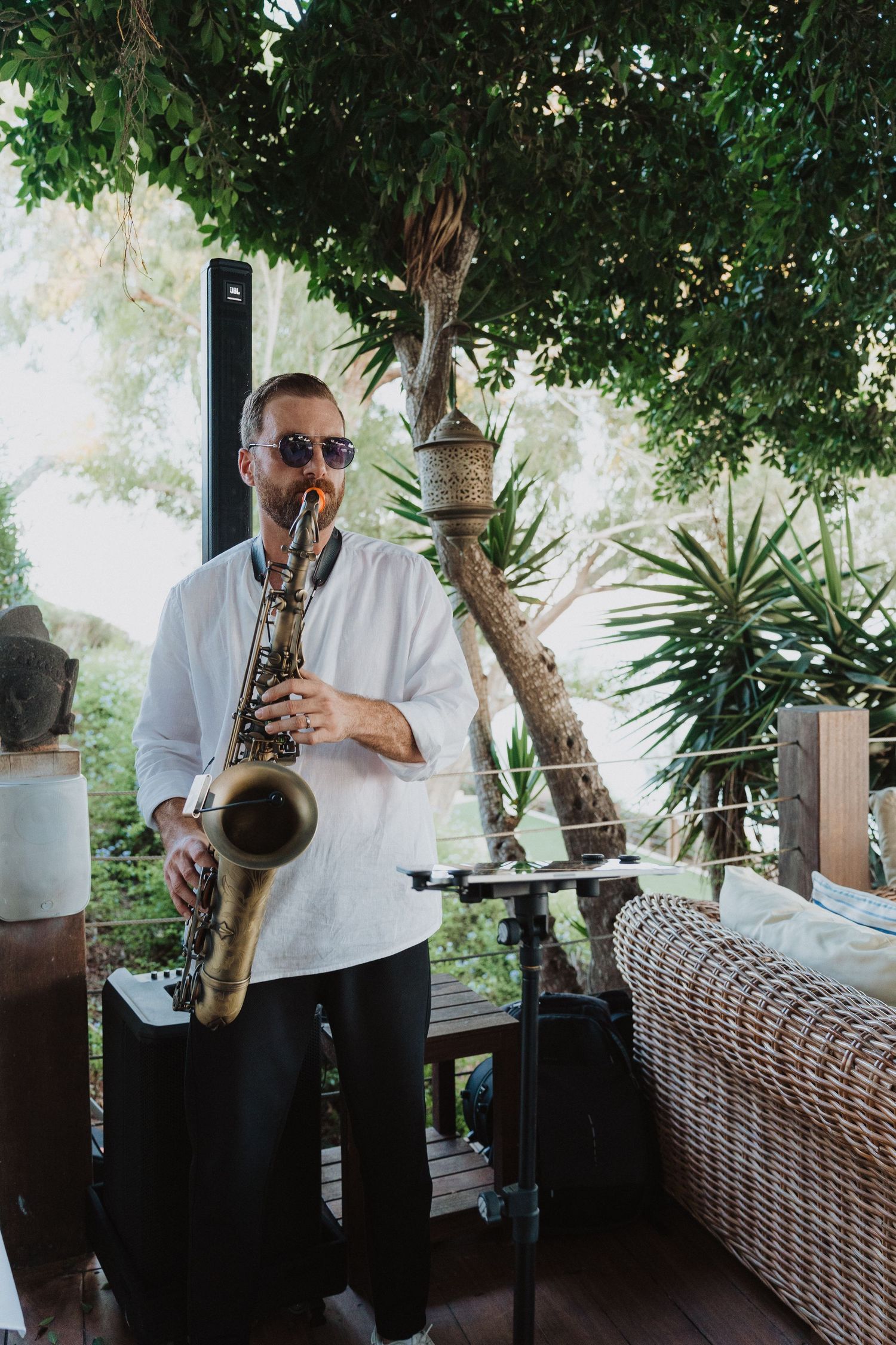 Saxophone player performing at an outdoor venue under tropical greenery.
