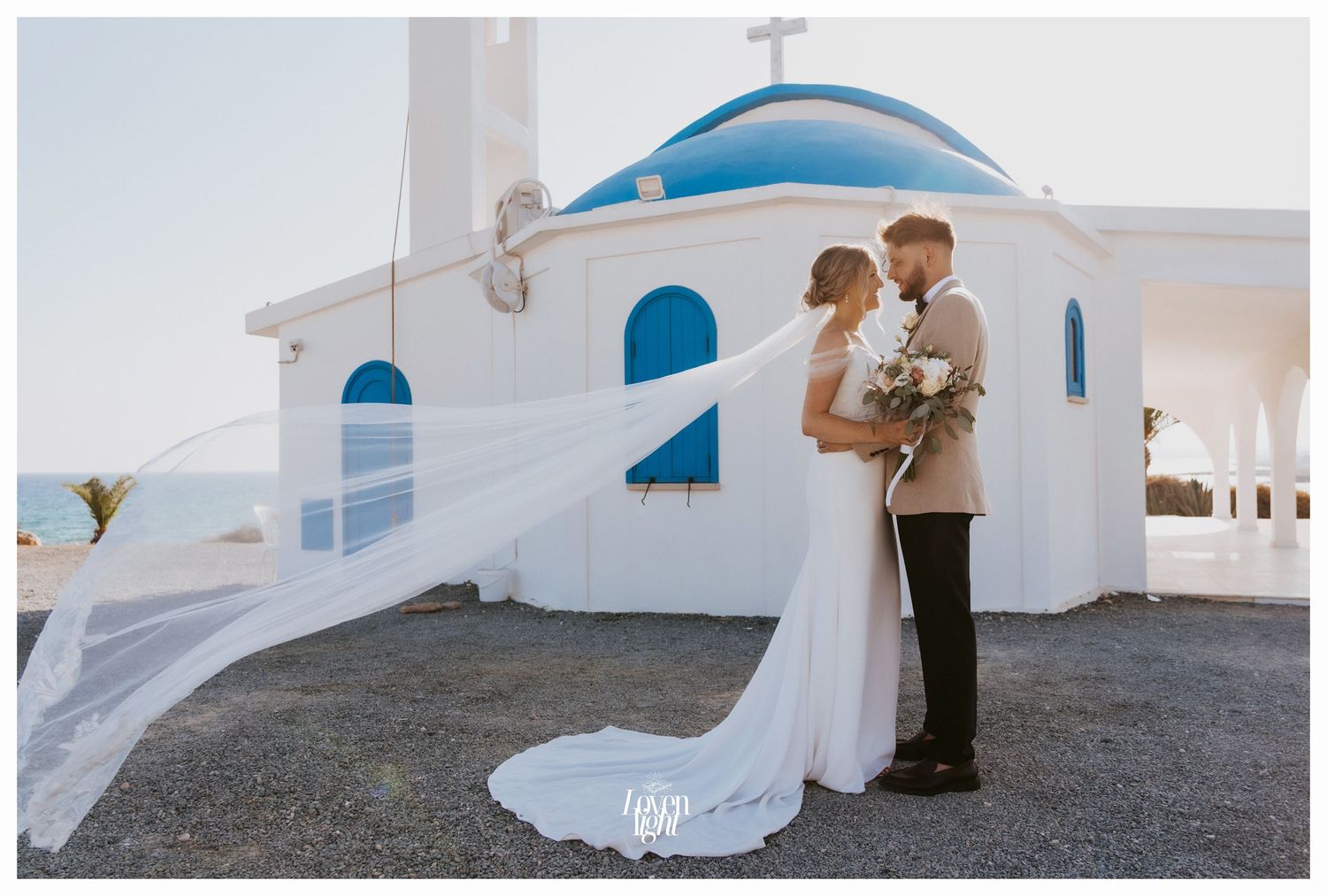 Wedding couple embraces in front of white Greek church with blue dome and flowing veil in Mediterranean setting.
