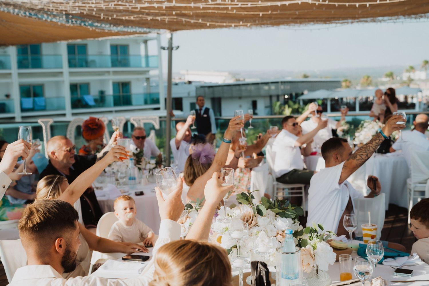 Wedding guests raise their glasses in a toast during an outdoor celebration by the sea.