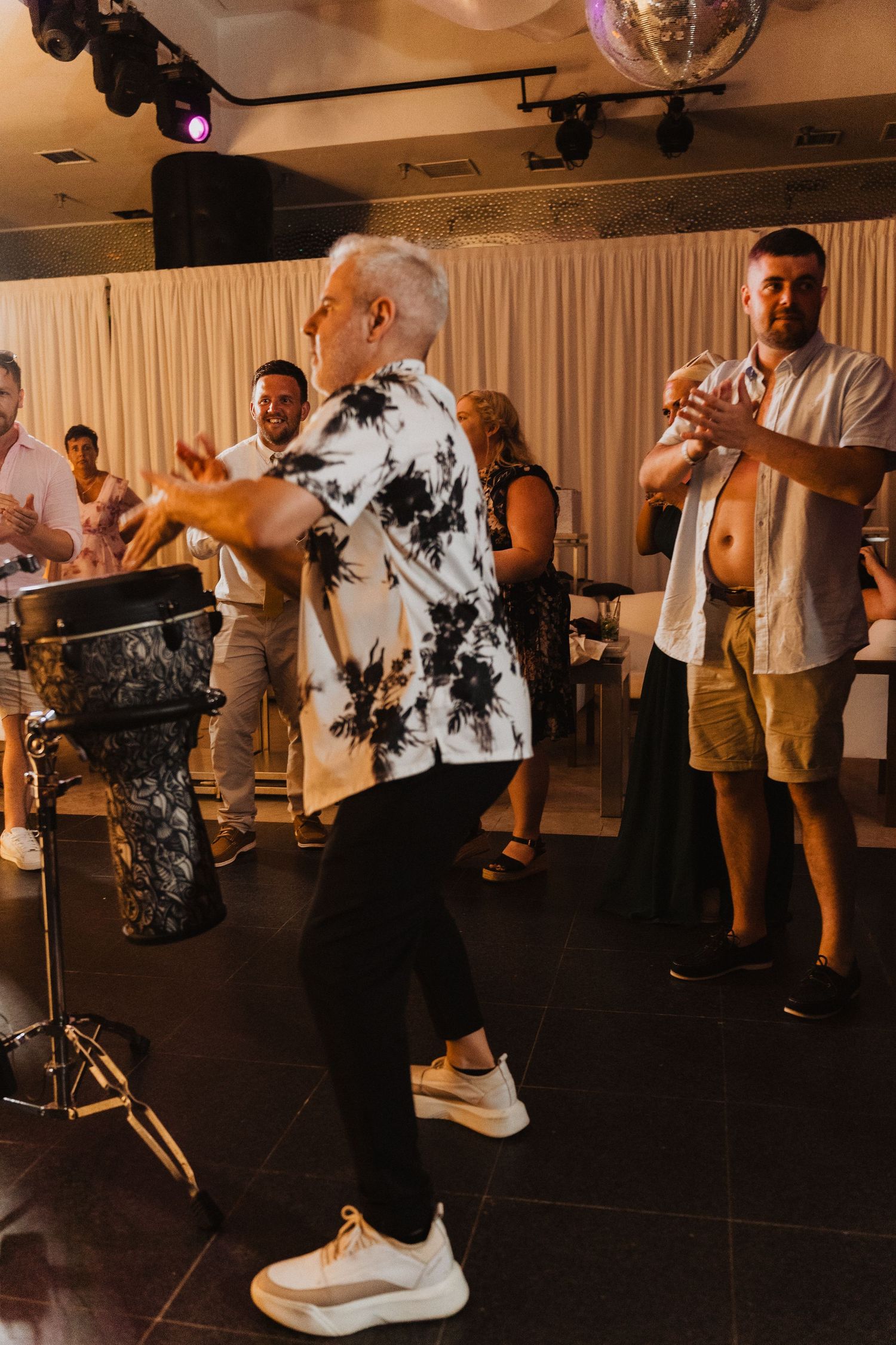 Wedding guests dancing and celebrating with percussion drum at indoor reception venue with curtained backdrop.
