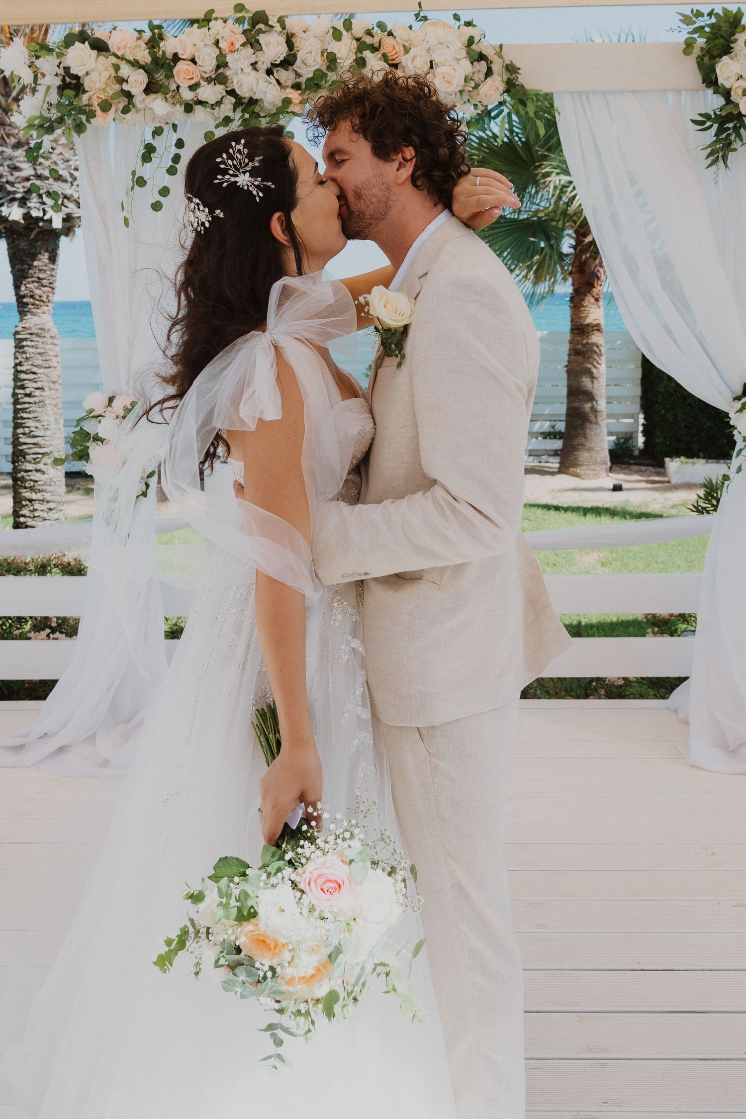 Newlyweds share intimate kiss under ornate floral arch with ocean view behind them.