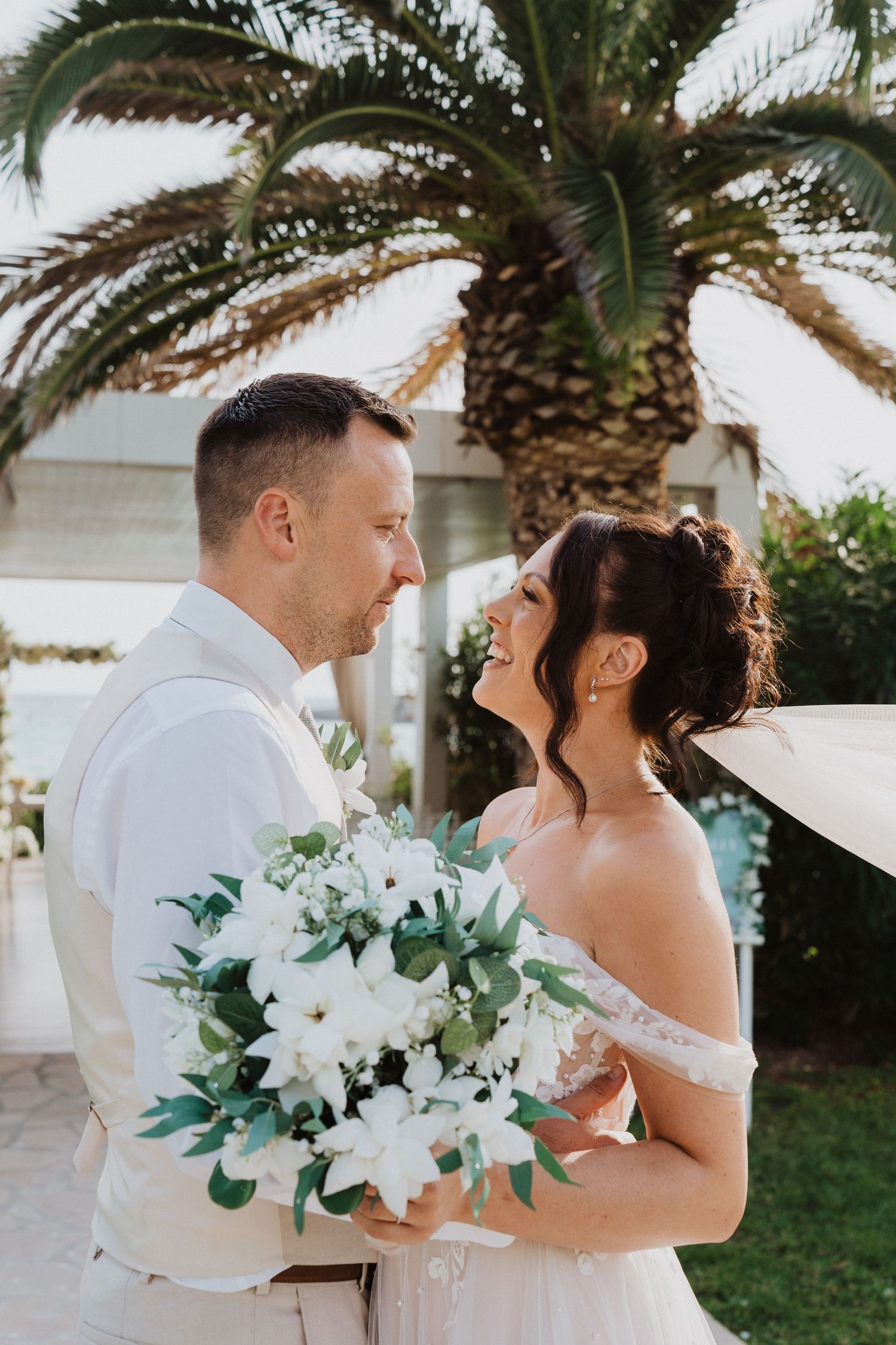 A romantic wedding couple poses with a beautiful white floral bouquet under a palm tree in a garden setting.