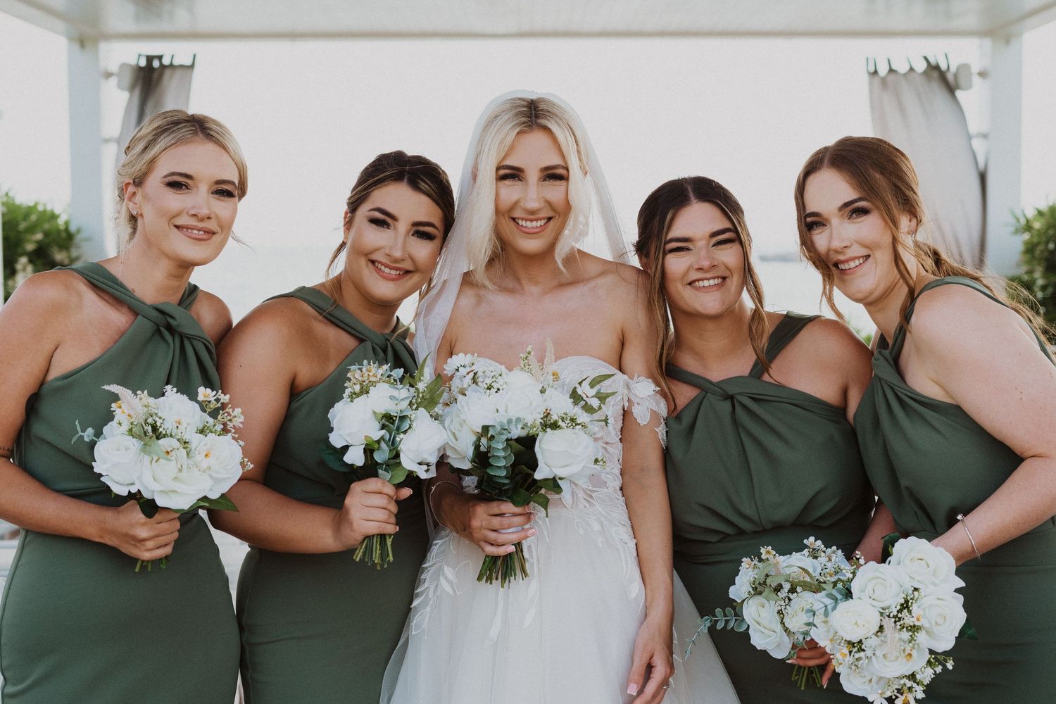 Bridal party wearing sage green dresses holds white floral bouquets under covered venue.
