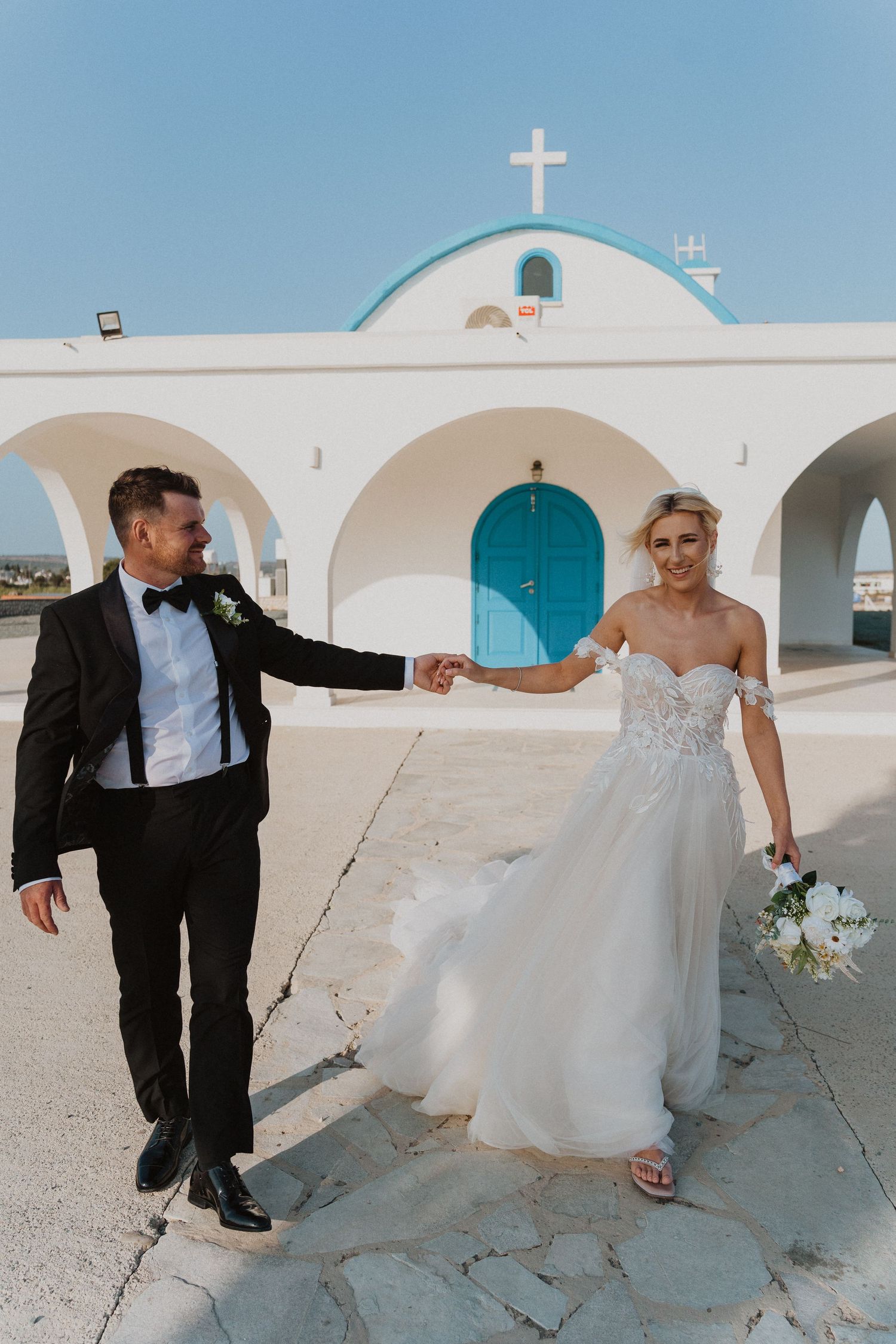 Elegant couple in wedding attire dances outside Greek orthodox chapel with blue door.