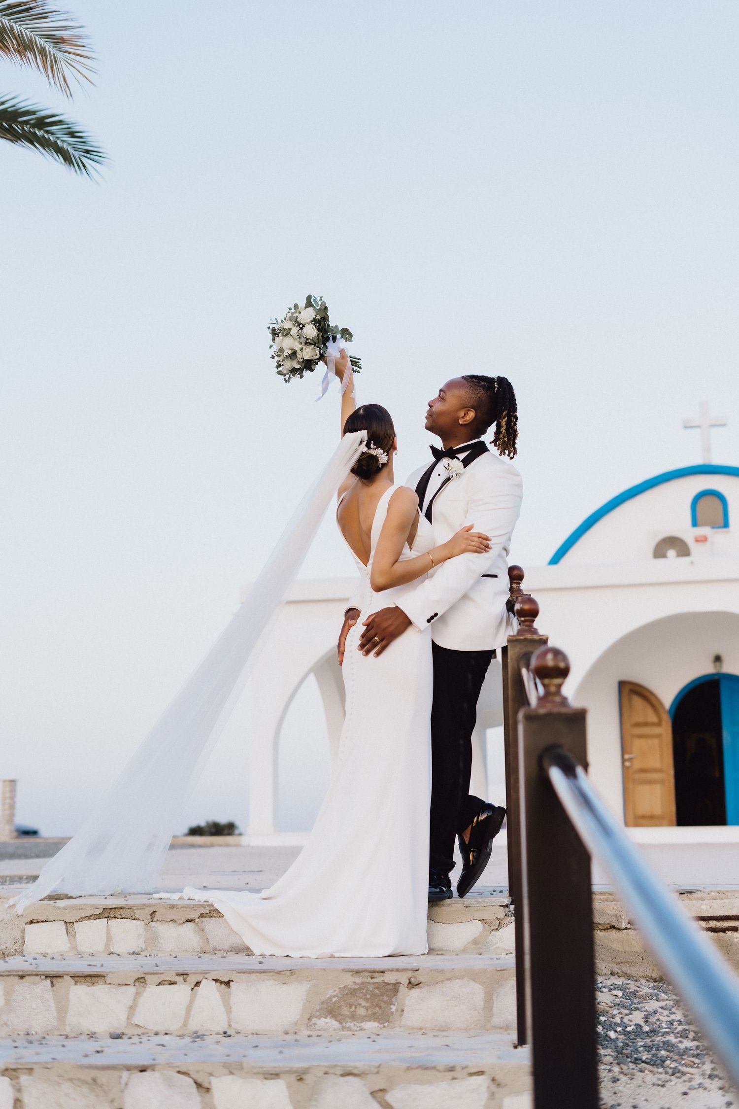 Wedding couple celebrating with raised bouquet on steps of a Mediterranean church with blue dome.