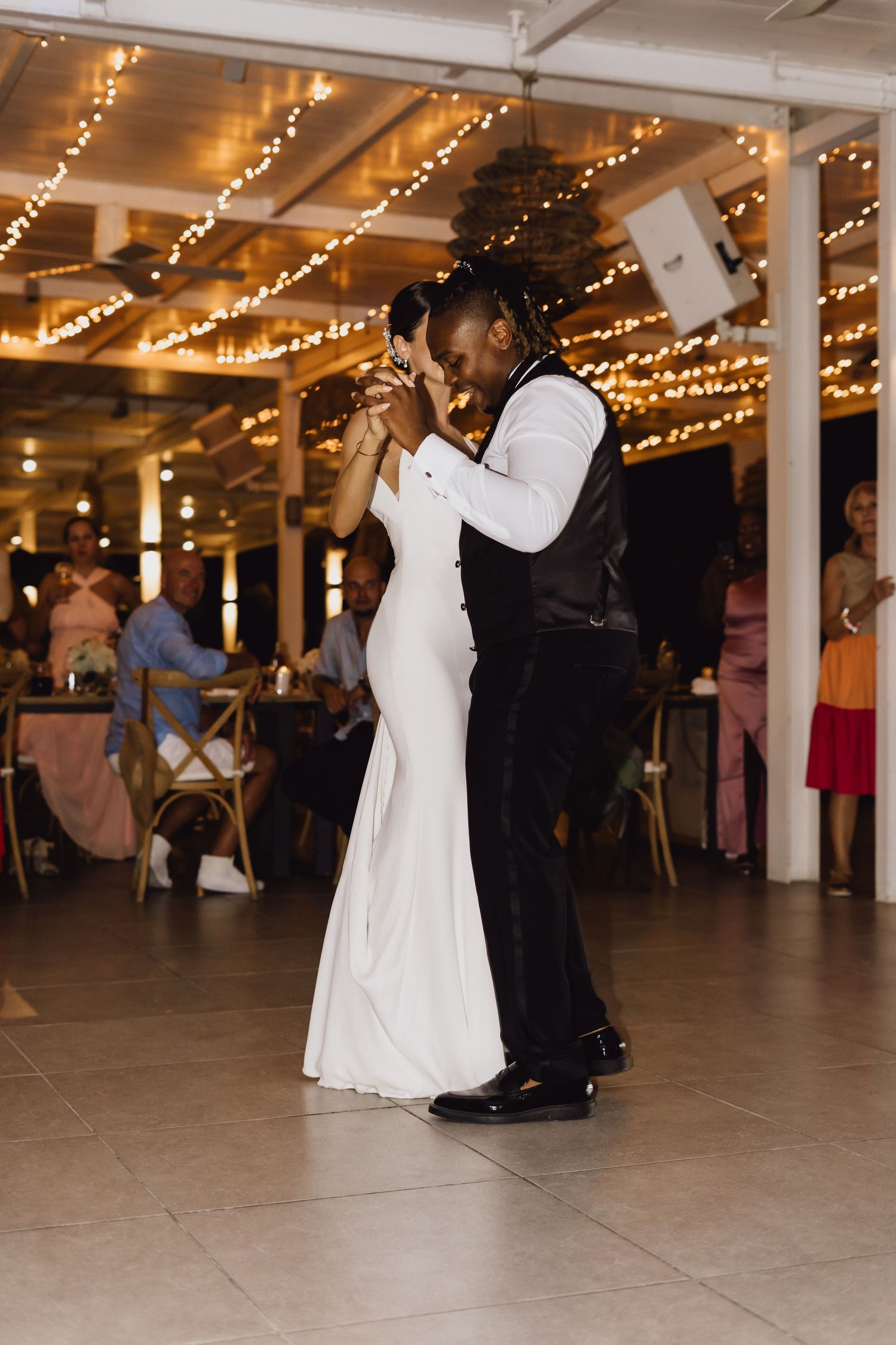 Wedding couple dancing together under warm string lights at indoor reception venue.