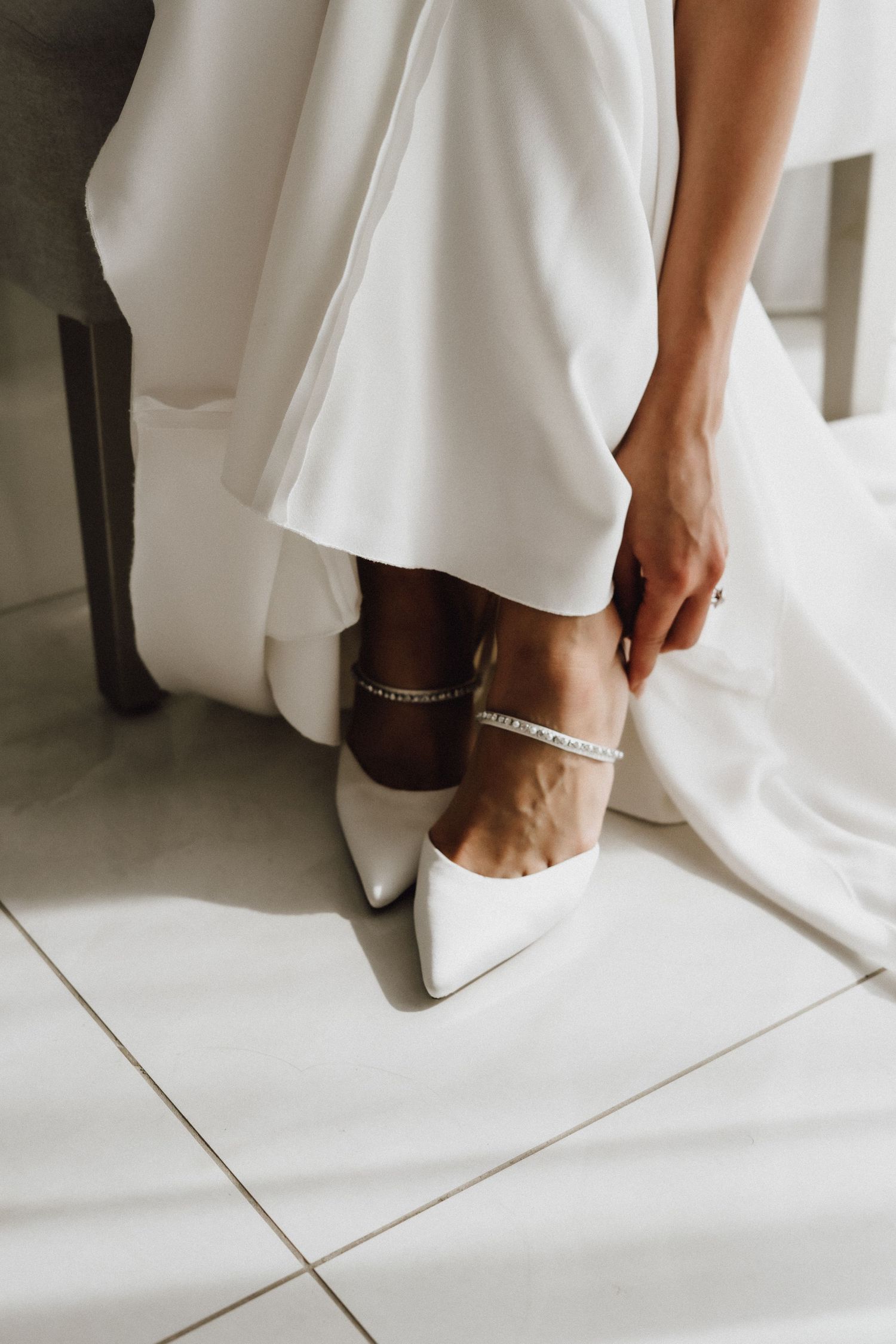 Close-up of white bridal shoes with delicate ankle straps against tile flooring.