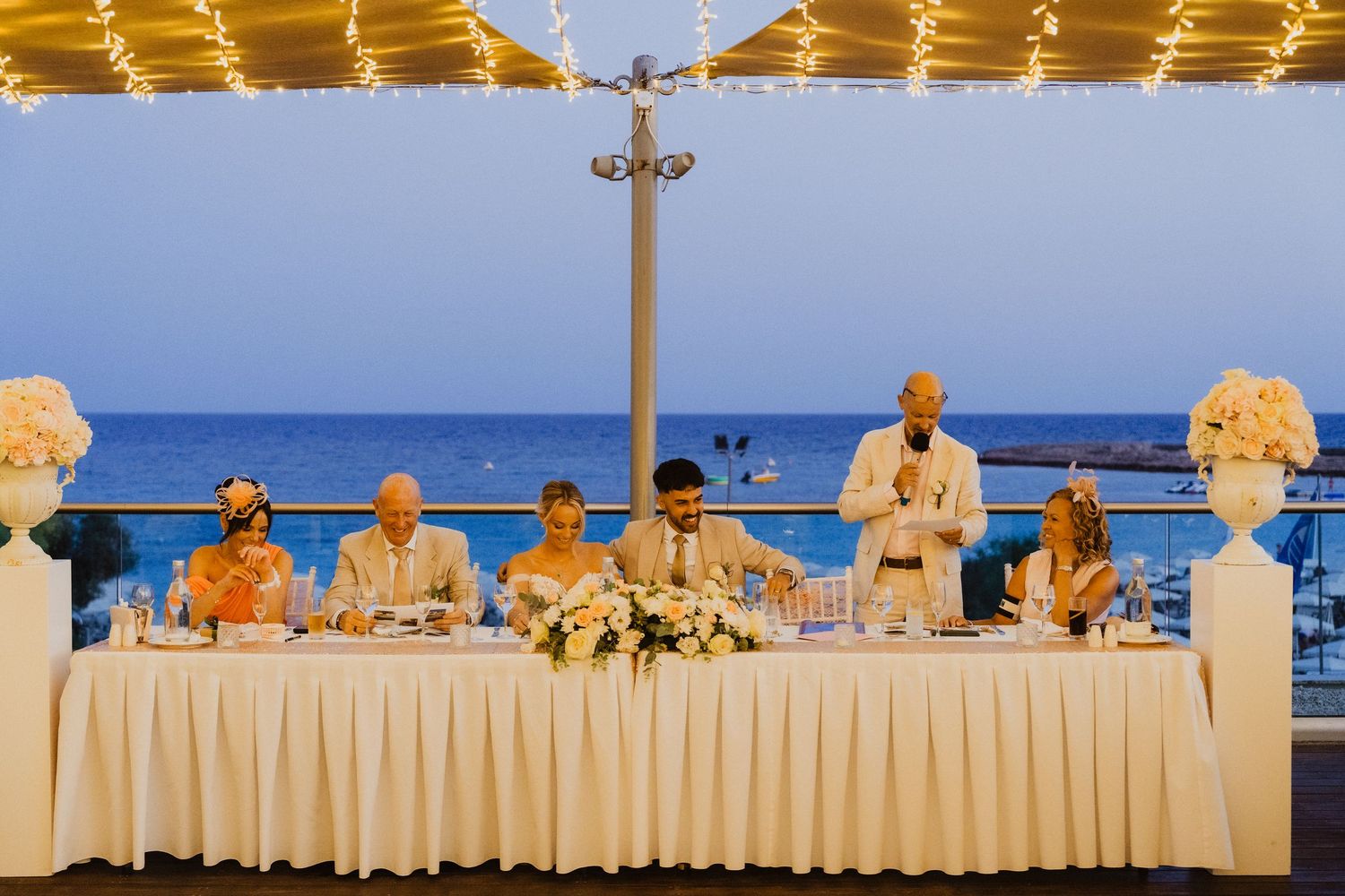 Wedding reception head table decorated with floral arrangements overlooking the ocean at sunset.