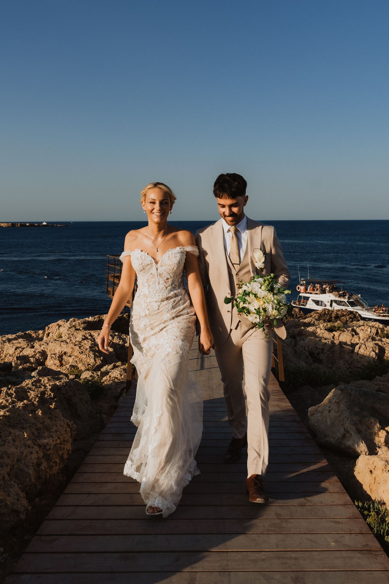 Couple walking along coastal boardwalk in wedding attire with ocean and rocky landscape.