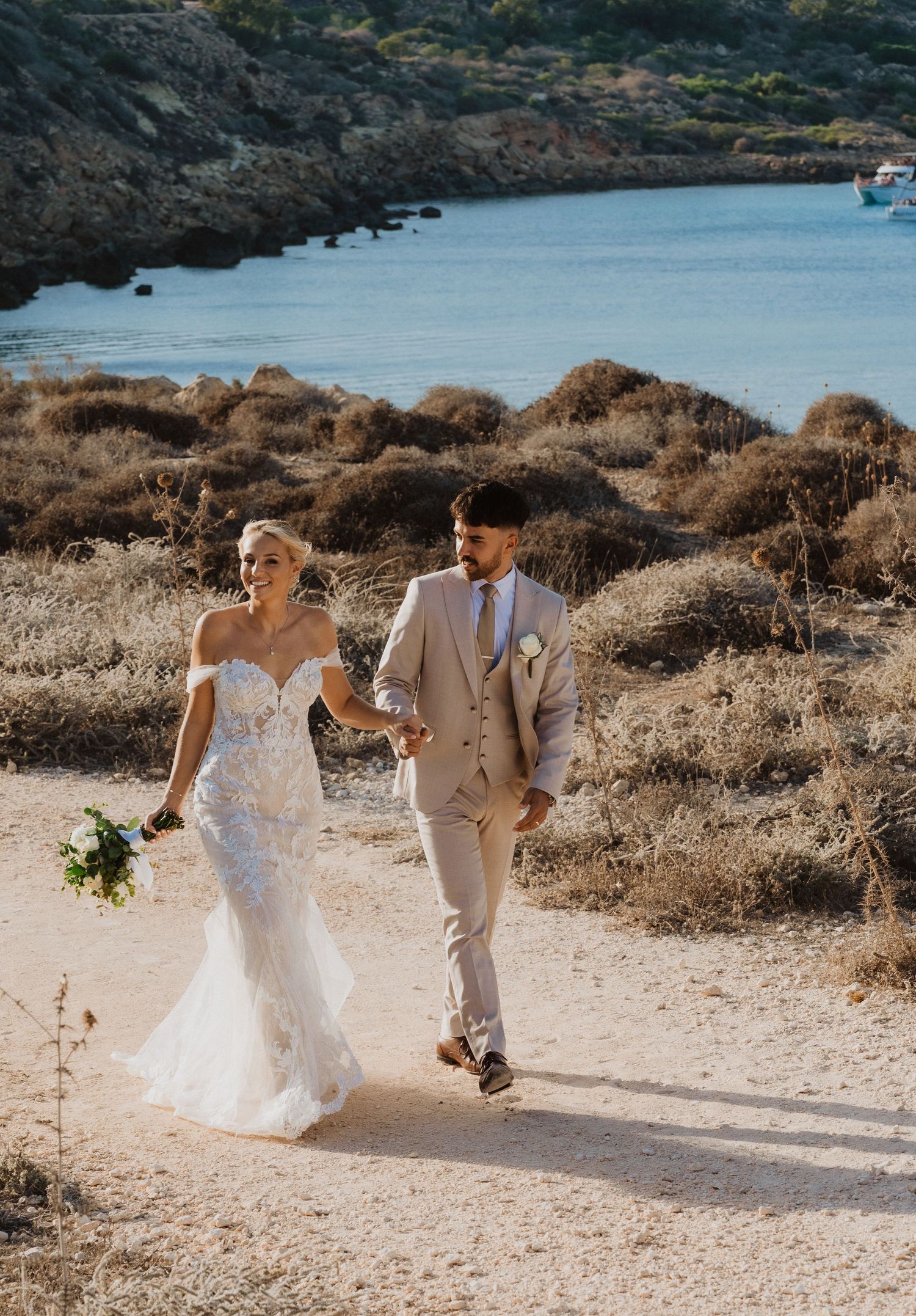 Newlyweds share a playful moment walking on a beach path with ocean and cliffs in the background.