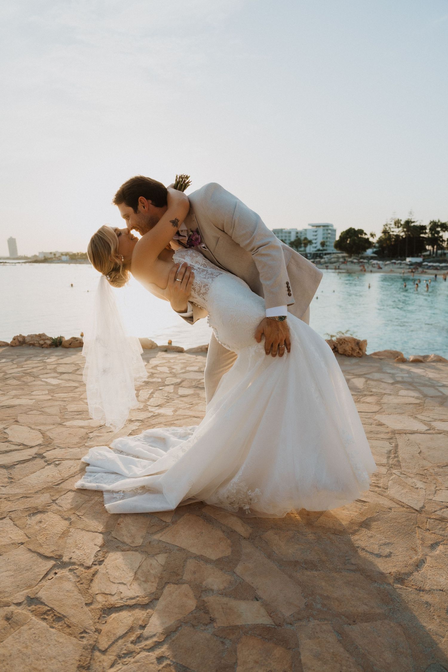 A white wedding dress flows elegantly across stone pavement near a waterfront setting during sunset.