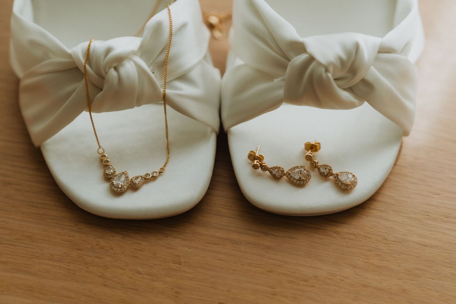 Elegant white bridal shoes with bow details and gold jewelry accents displayed on wooden surface.