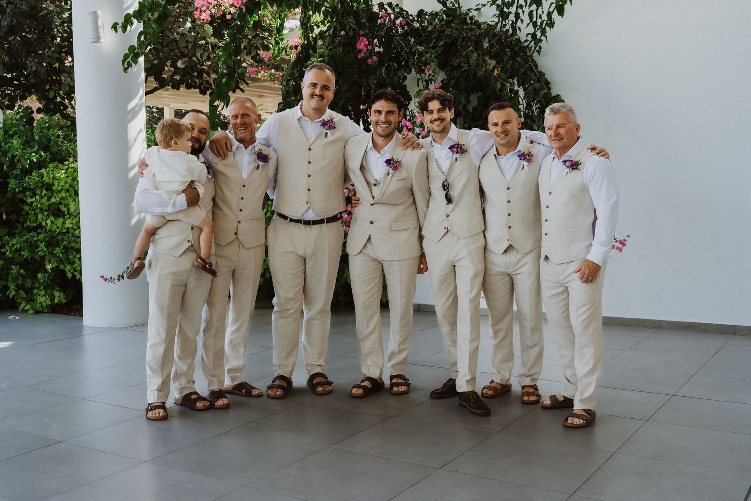 Groomsmen in matching cream suits pose casually along white resort walkway.