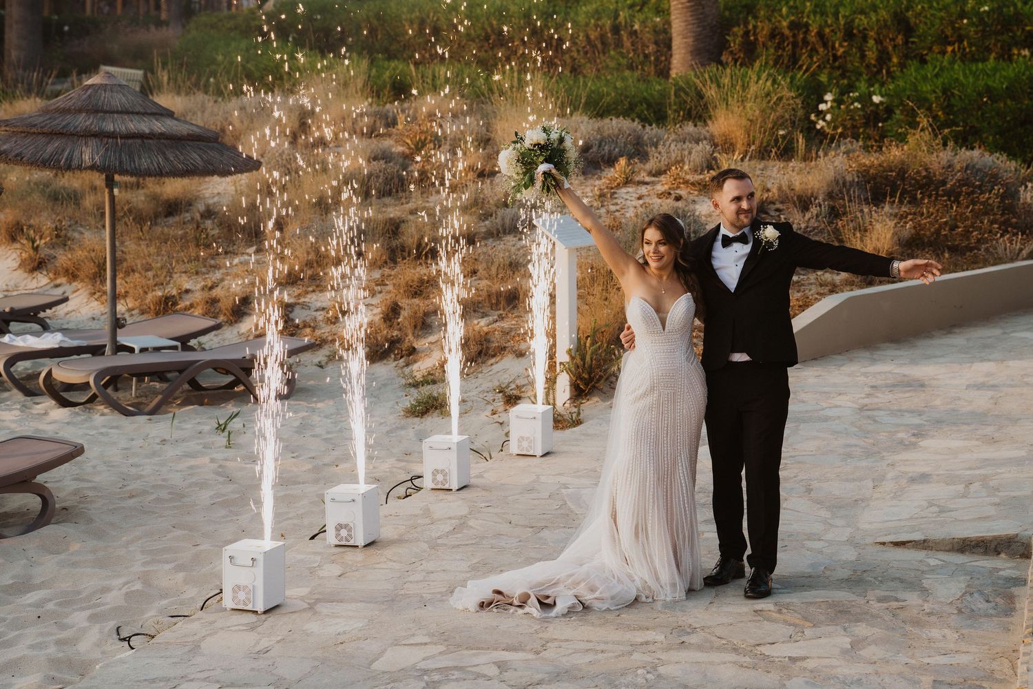 Bride and groom celebrate with sparkling pyrotechnic fountains on a sandy beach at sunset.