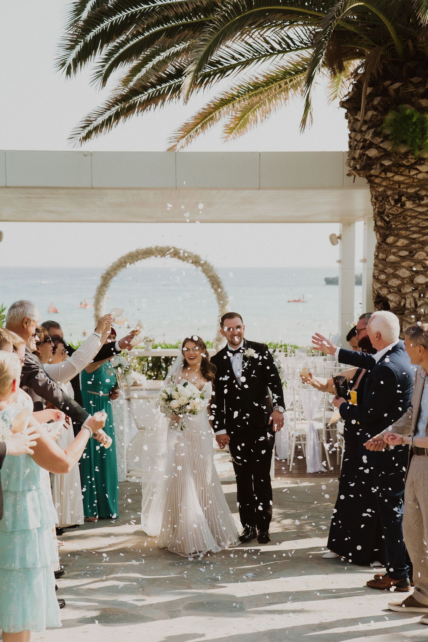 Bride and groom walk down the aisle as guests toss white confetti at an outdoor seaside wedding ceremony.