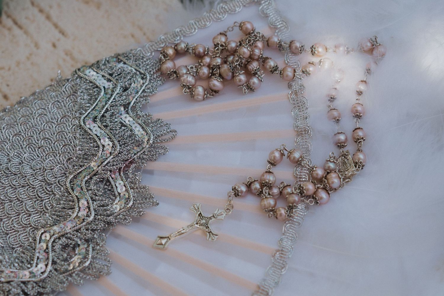 Vintage pink pearl rosary beads with gold crucifix resting on silver lace fan and white fabric background.