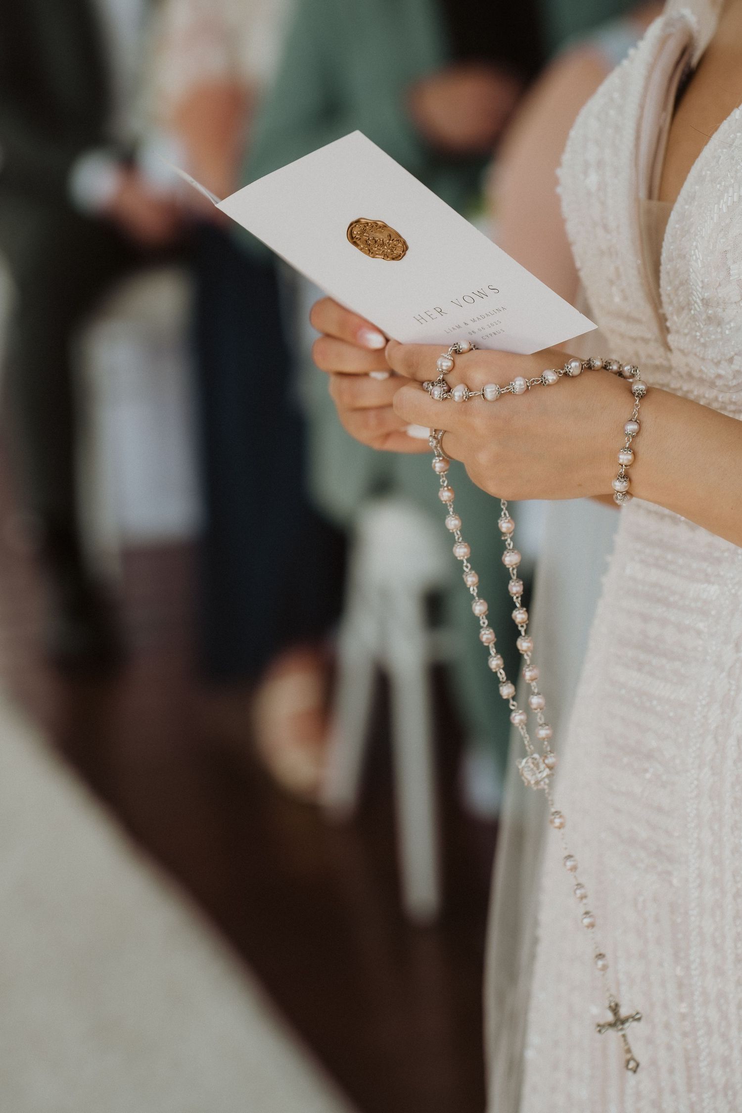 Bride holding rosary beads and ceremony booklet with gold wax seal during wedding.