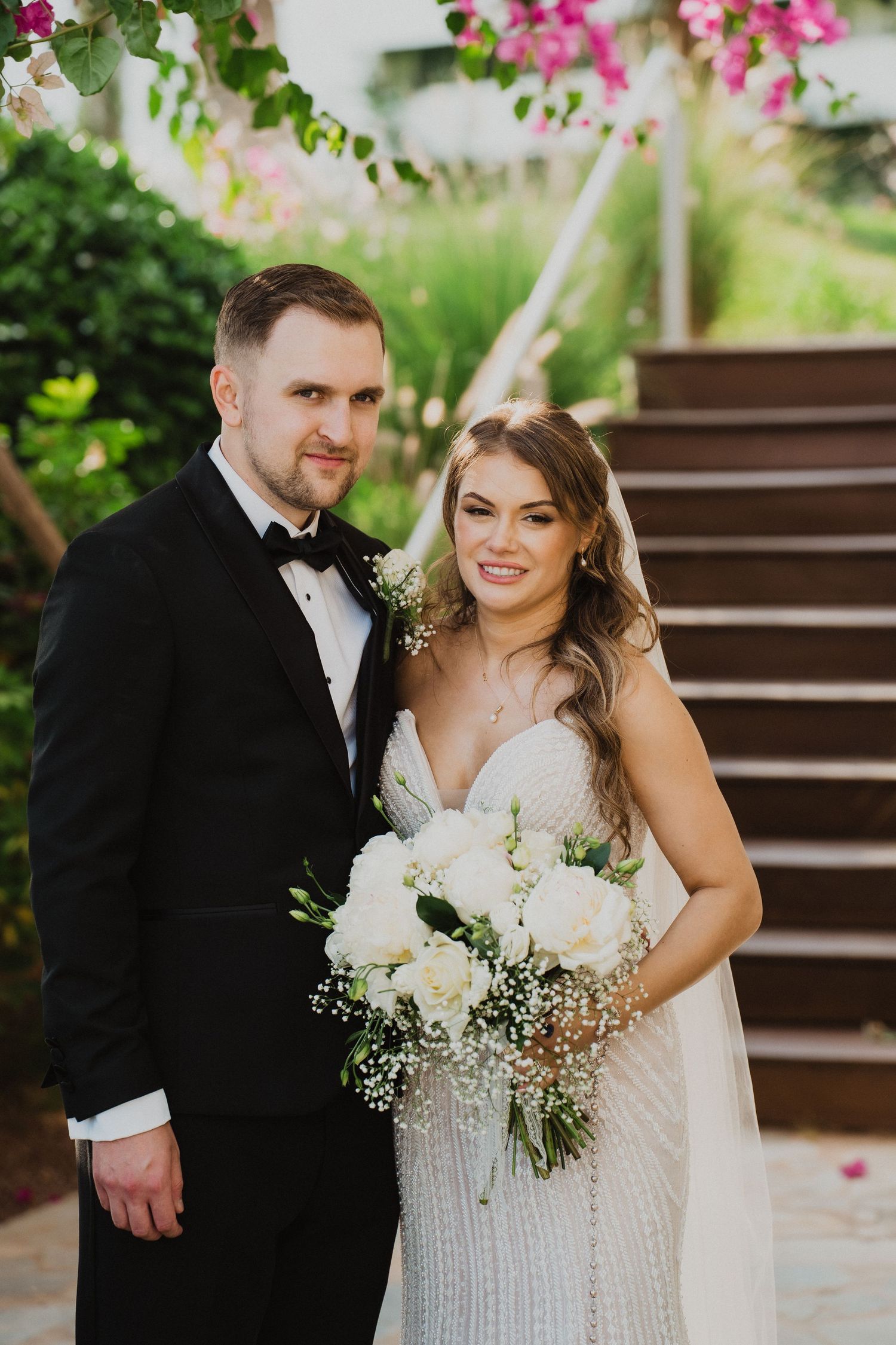 Smiling bride holding white rose bouquet stands with groom in black tuxedo at an outdoor wedding venue.