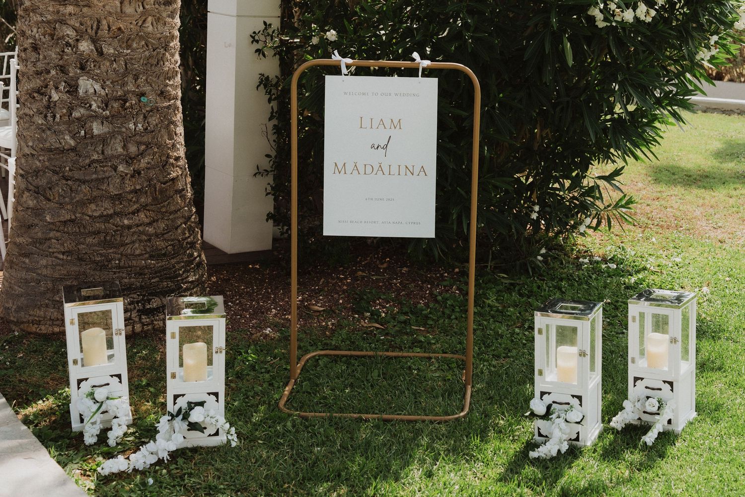 Outdoor wedding welcome sign for Liam and Madalina on gold stand with white lantern decorations.