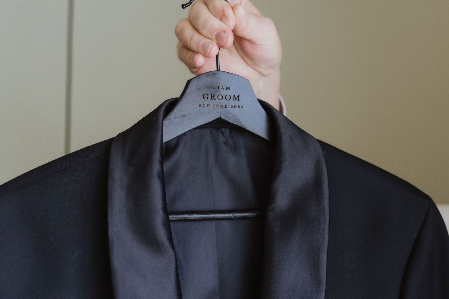 Black groom suit jacket on a personalized wedding hanger held by hand against neutral background.