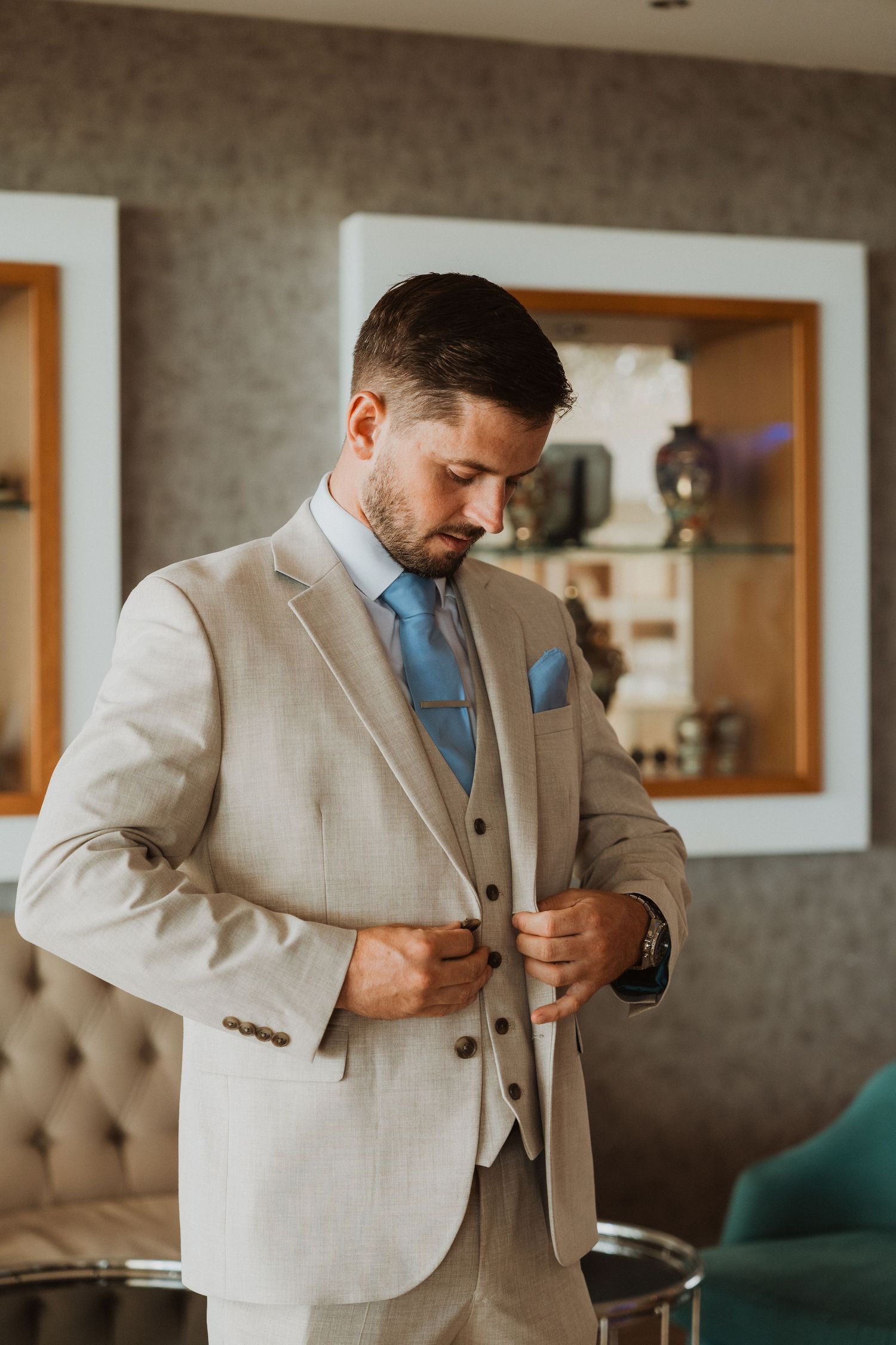 A well-dressed groom in a beige three-piece suit with blue tie adjusts his jacket button indoors.