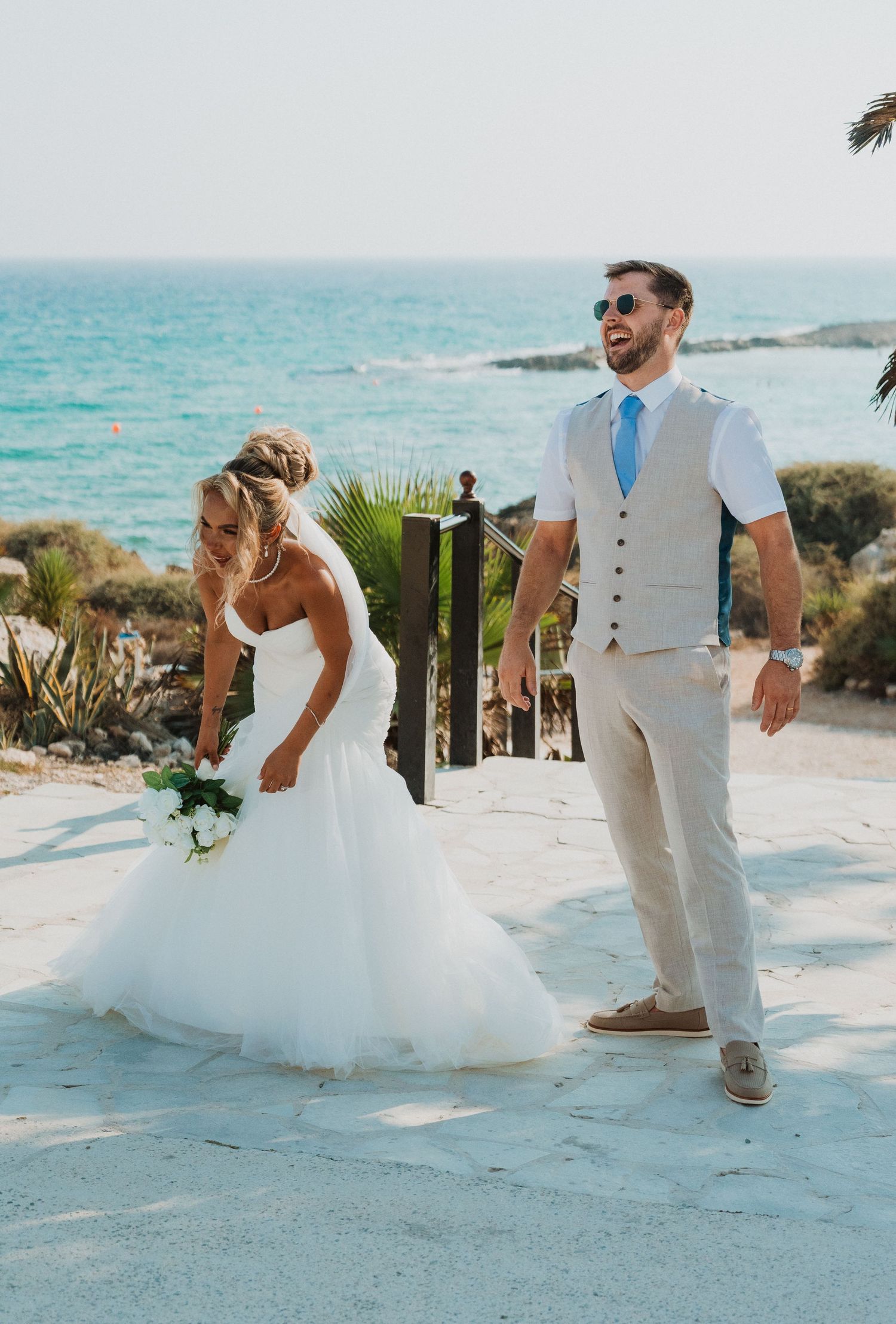 Bride in white gown and groom in tan suit celebrate beach wedding with ocean views.