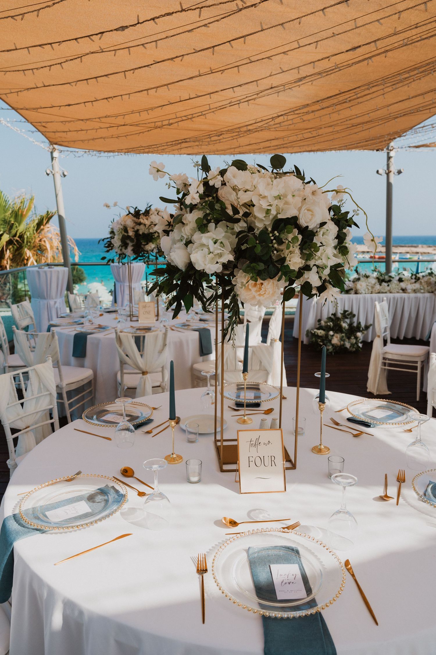 Elegant beach wedding reception table with white floral centerpiece, gold cutlery, blue napkins, and turquoise ocean view.