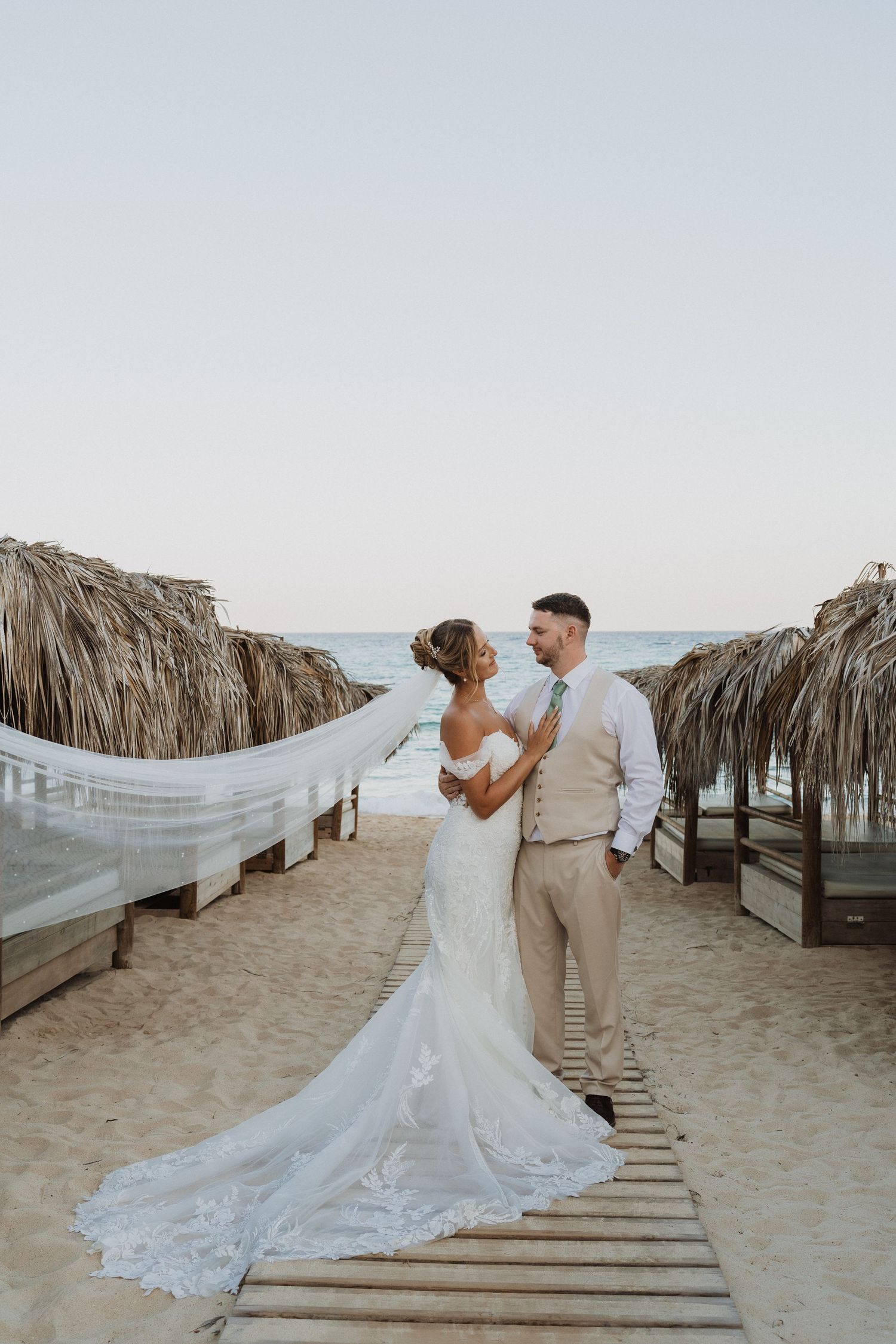 A romantic beach wedding photoshoot with a couple on a wooden boardwalk surrounded by thatched umbrellas at sunset.