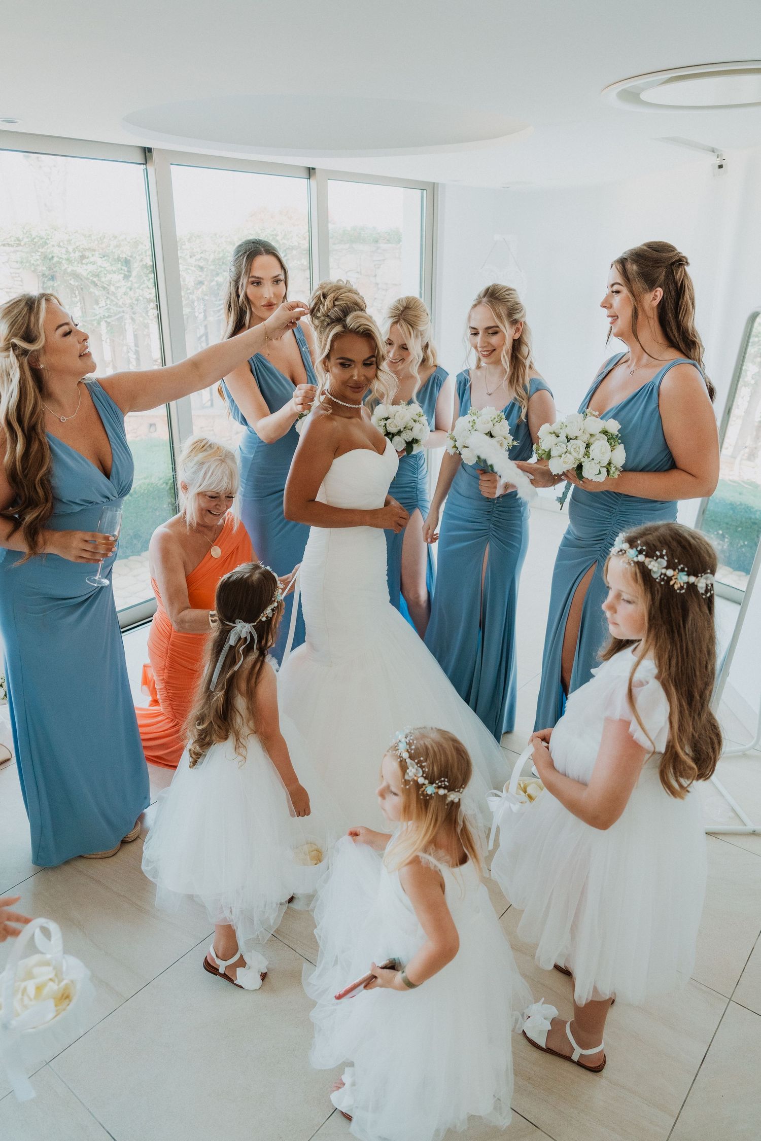 Bride in white gown surrounded by bridesmaids in blue dresses and flower girls in white, preparing for wedding ceremony.