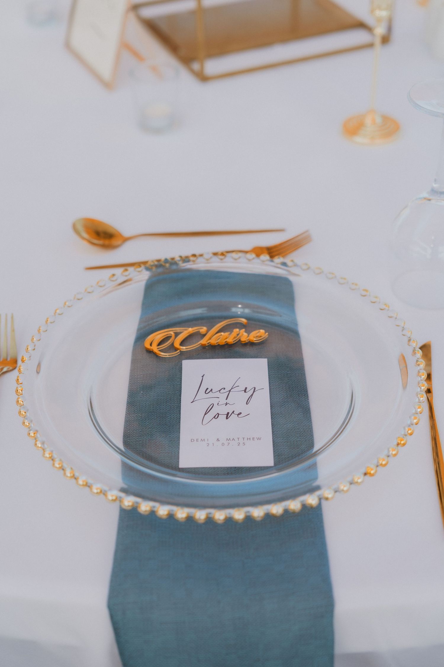 Elegant wedding place setting with gold beaded charger, blue napkin, name card Claire, and Lucky in Love menu card.