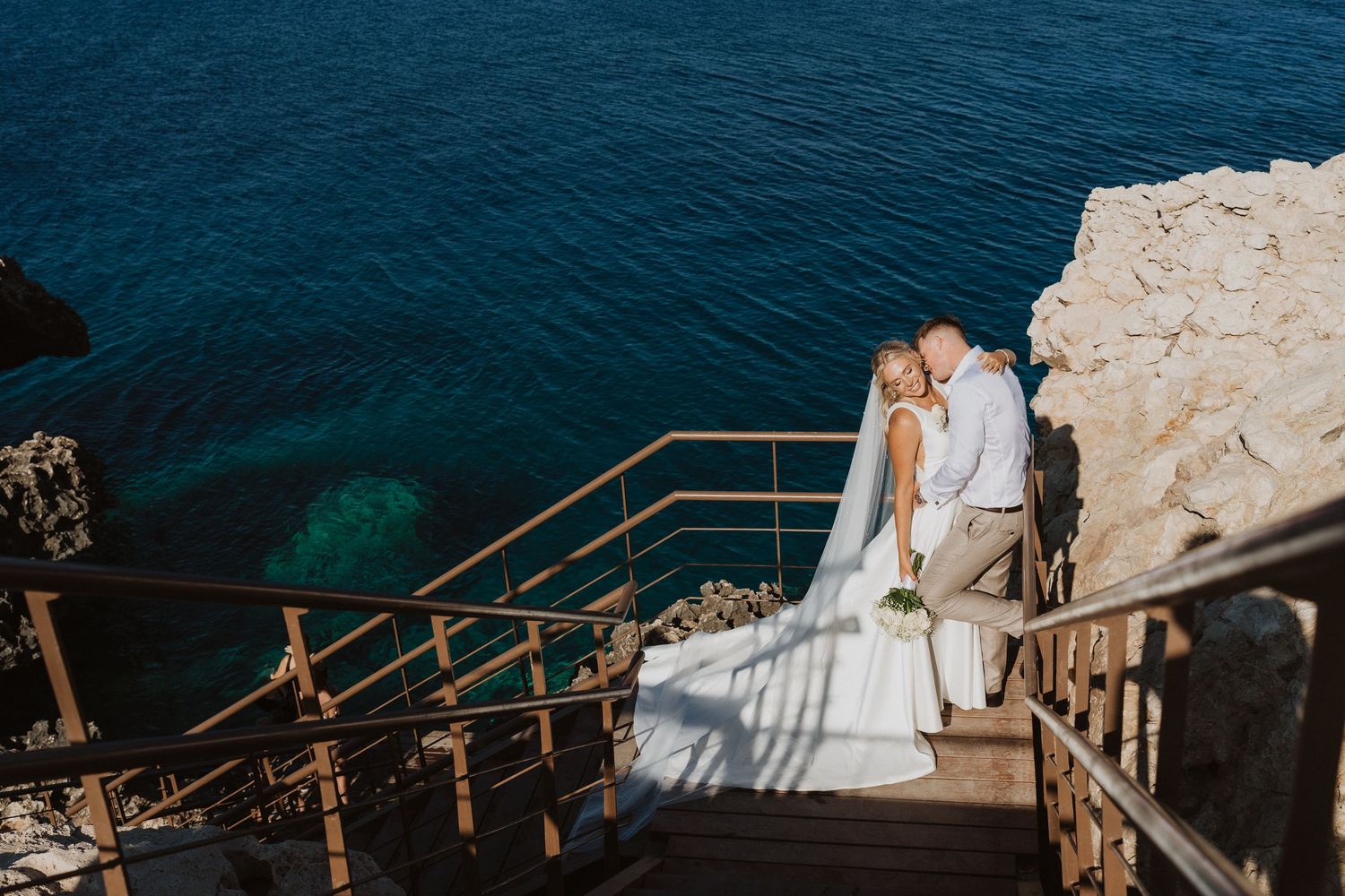 A couple in wedding attire walks down wooden steps leading to the Mediterranean Sea on a sunny day.