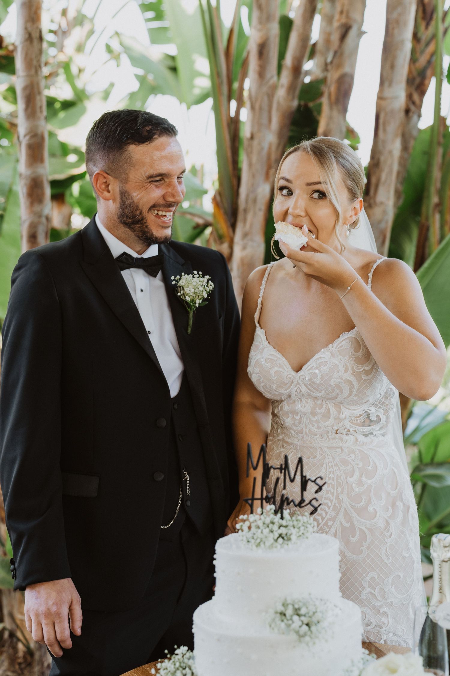 A joyful wedding couple shares a candid moment while cutting their white wedding cake adorned with flowers.