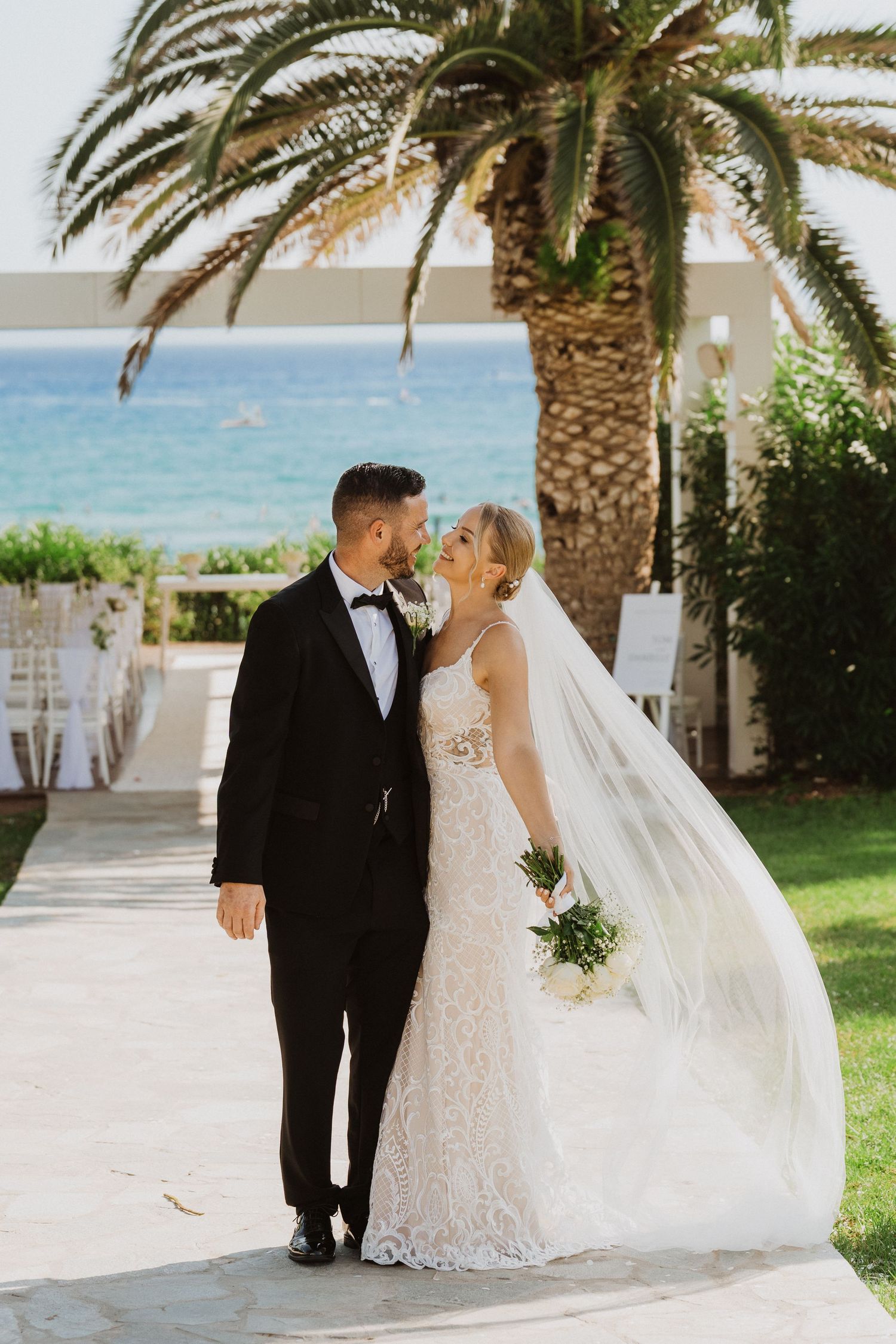 A romantic wedding couple shares a tender moment under palm trees at a seaside venue with a flowing white veil.